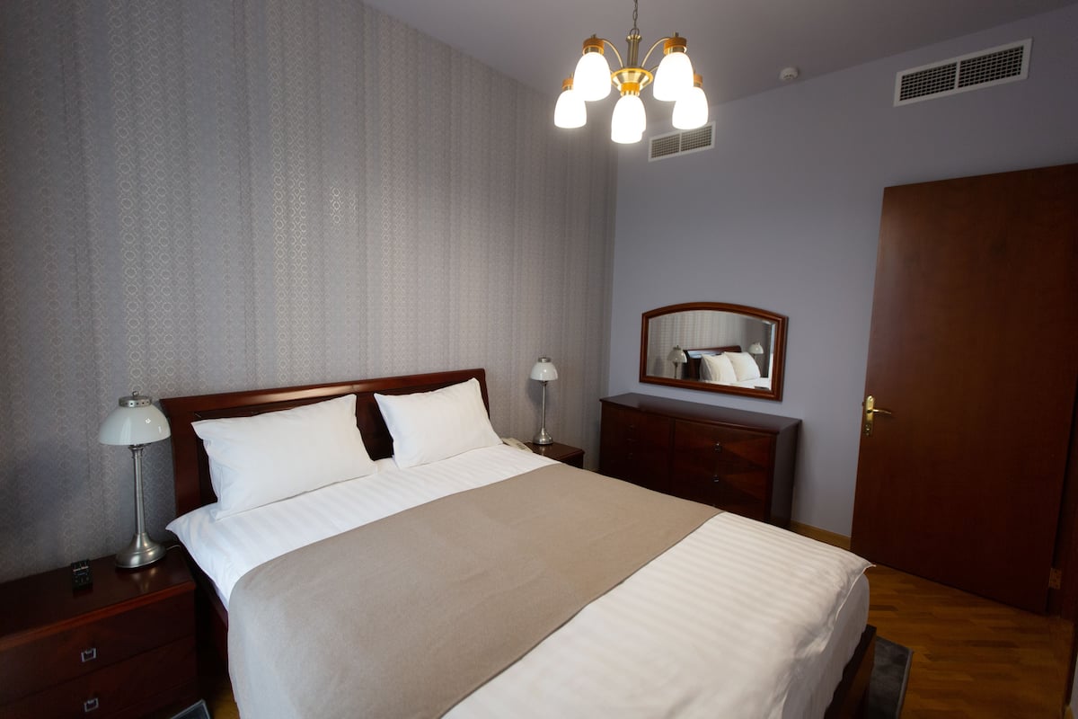 Uma cama bem arrumada com cabeceira de madeira, luminária de cabeceira, mesa de apoio, cômoda e um espelho na parede.