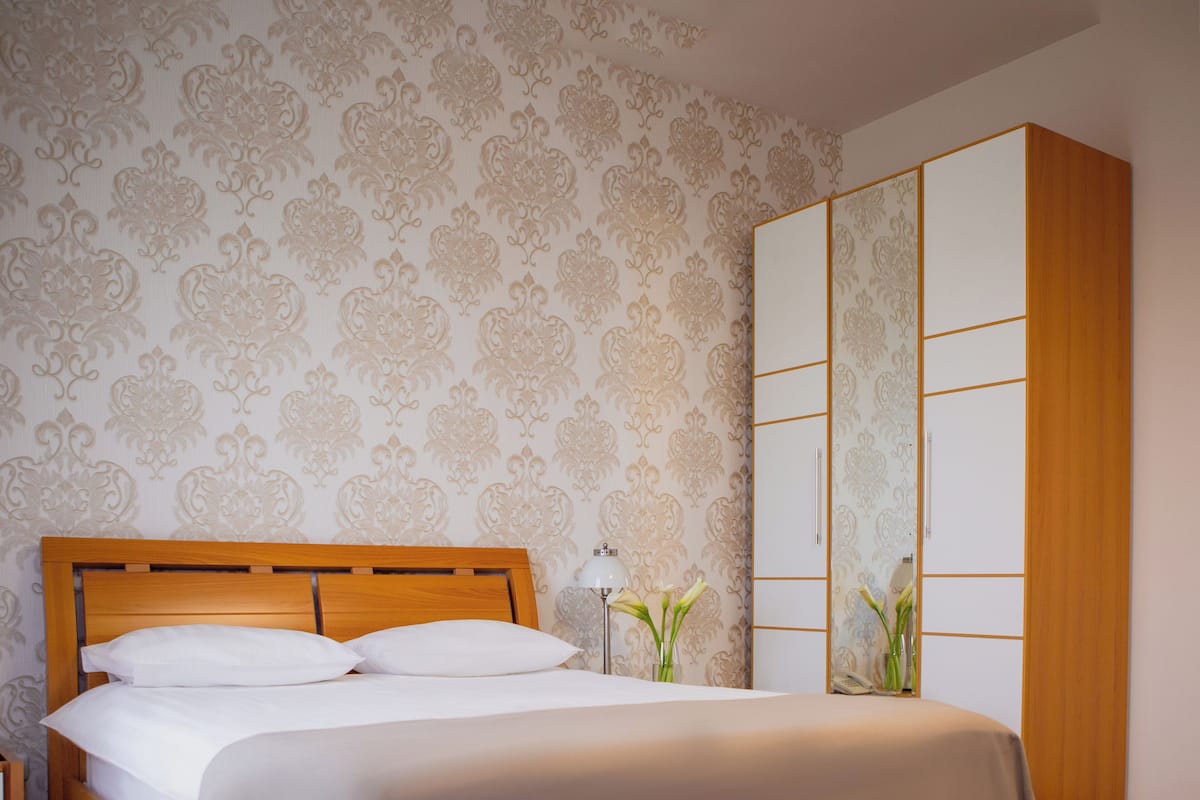 Cama bem arrumada com roupa de cama branca em quarto com papel de parede estampado e guarda-roupa de madeira.