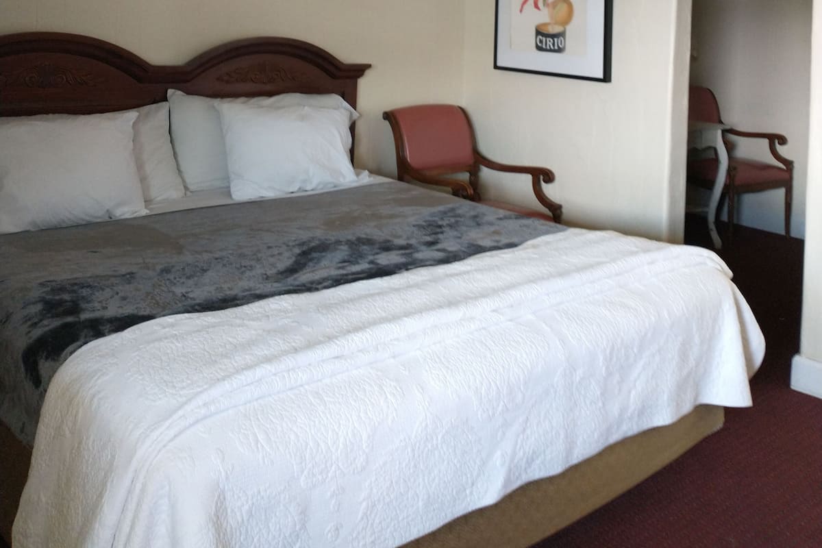 Quarto de hotel com uma cama grande, uma cadeira, um quadro na parede e uma porta que dá para outro cômodo.