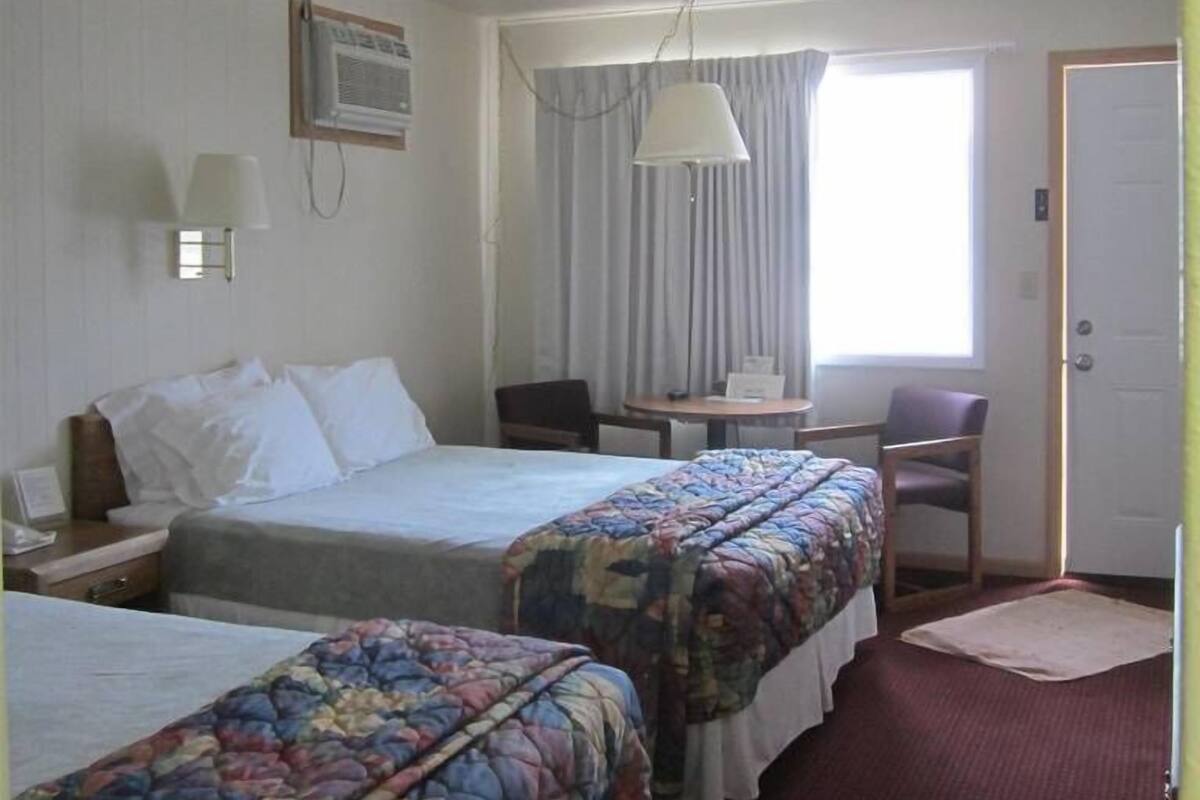 Quarto de hotel com duas camas, uma escrivaninha, uma cadeira e uma janela com cortinas.