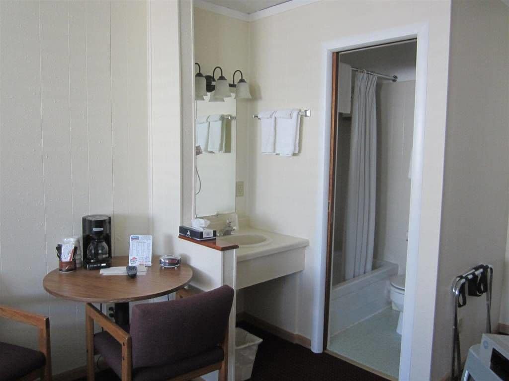 A hotel room with a round table, a chair, a coffee maker, a sink, a mirror, and a shower area.
