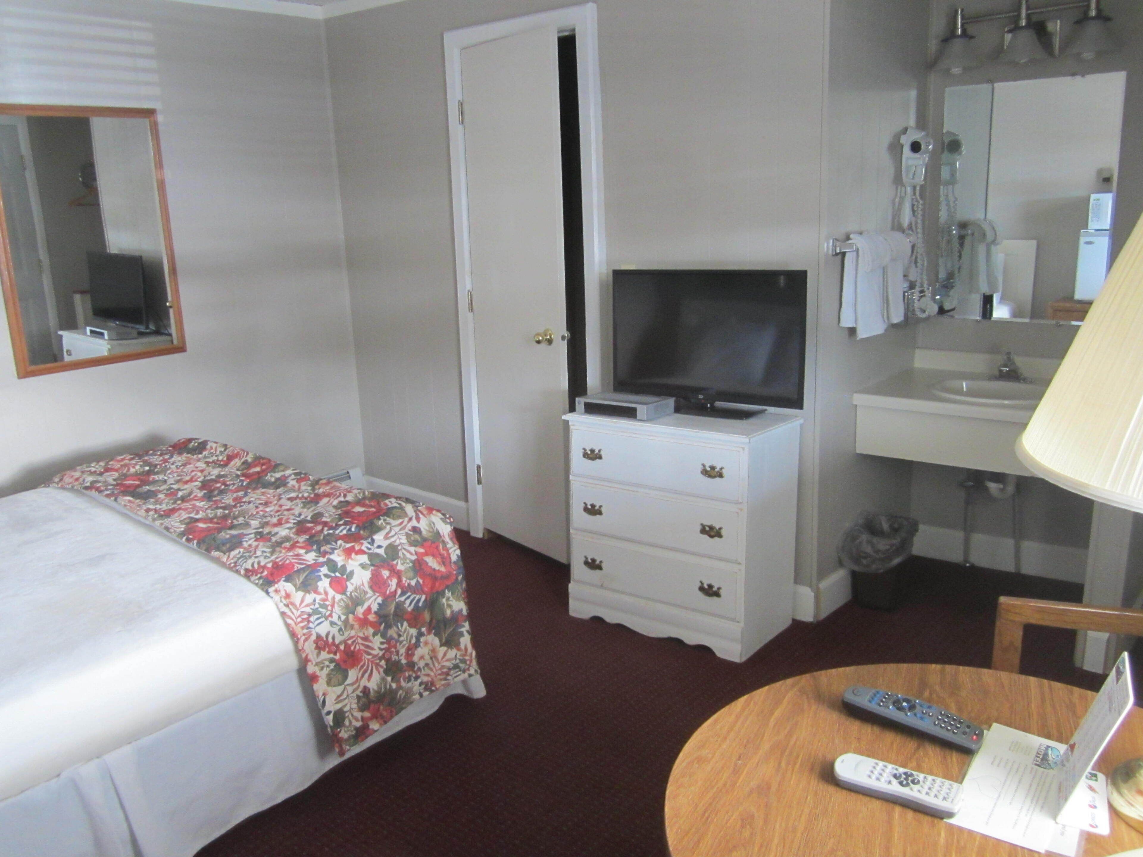 Ein Hotelzimmer mit einem Bett, einem Schminktisch mit Fernseher, einem Badezimmer mit Waschbecken und Spiegel sowie einem runden Tisch mit Fernbedienung.