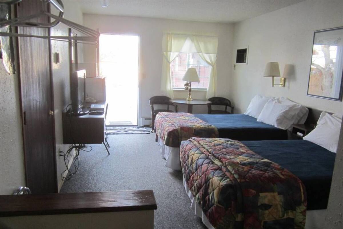 Quarto de hotel com duas camas, uma escrivaninha, uma cadeira, um abajur e uma janela com cortinas.