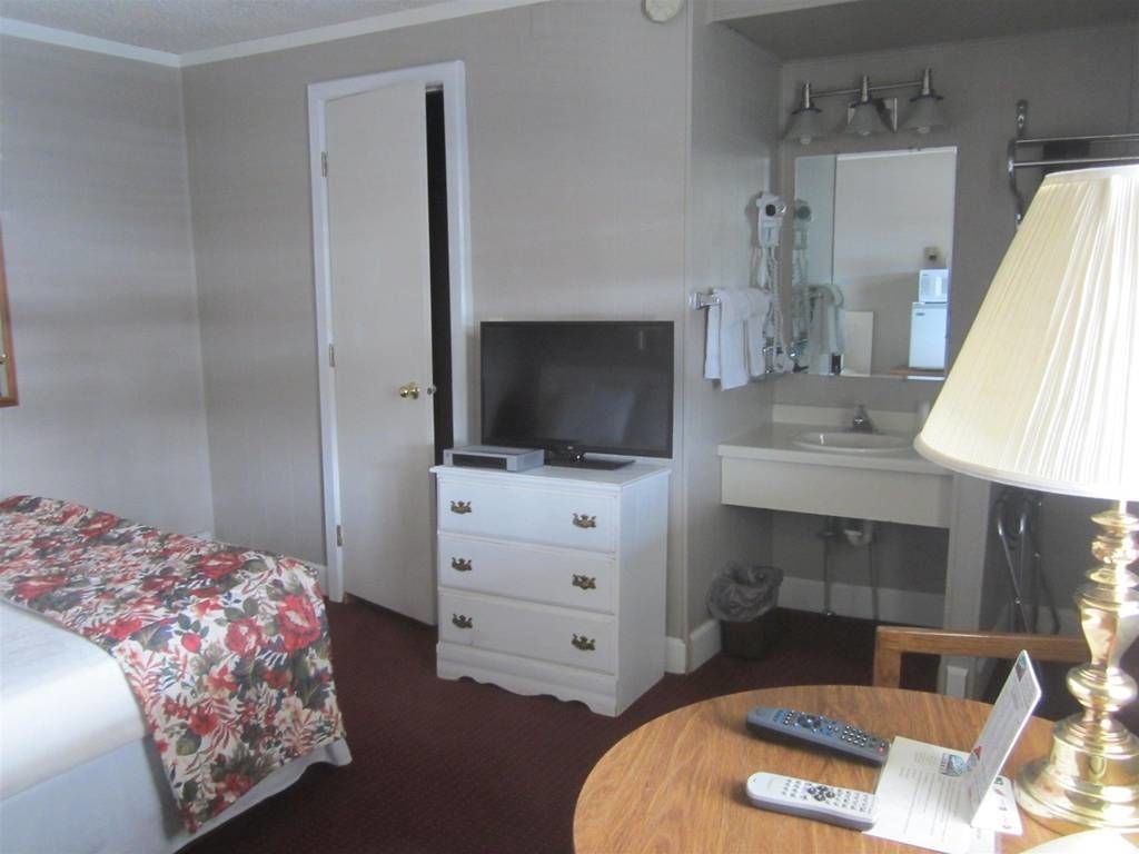 Ein Hotelzimmer mit Bett, Schminktisch, Fernseher, Bad mit Waschbecken und Spiegel sowie rundem Tisch mit Lampe und Fernbedienungen.