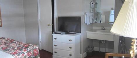 Ein Hotelzimmer mit Bett, Schminktisch, Fernseher, Bad mit Waschbecken und Spiegel sowie rundem Tisch mit Lampe und Fernbedienungen.