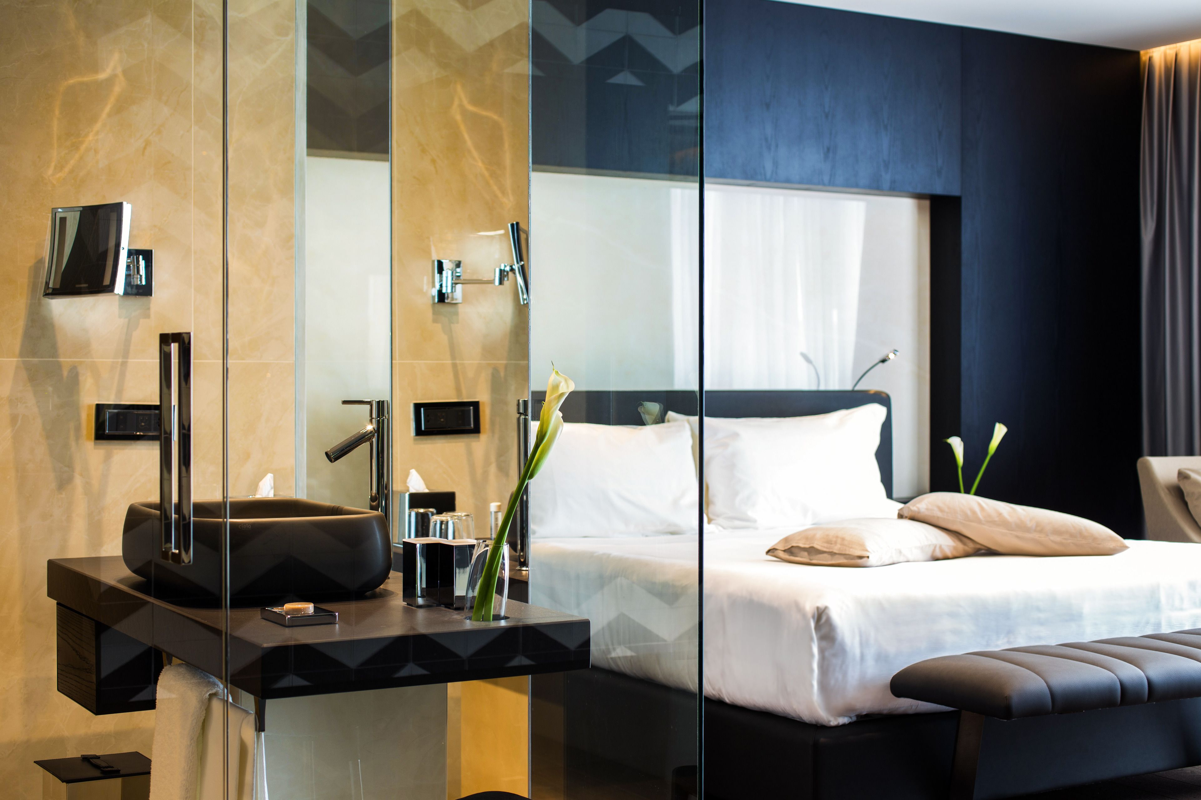 A modern hotel room with a glass-enclosed bathroom, a large bed with white linens, and a black leather bench.