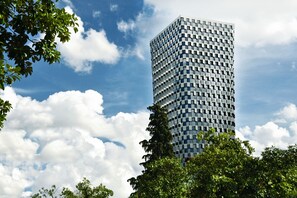 A tall, modern building with a grid-like facade, surrounded by lush green trees and a partly cloudy sky.