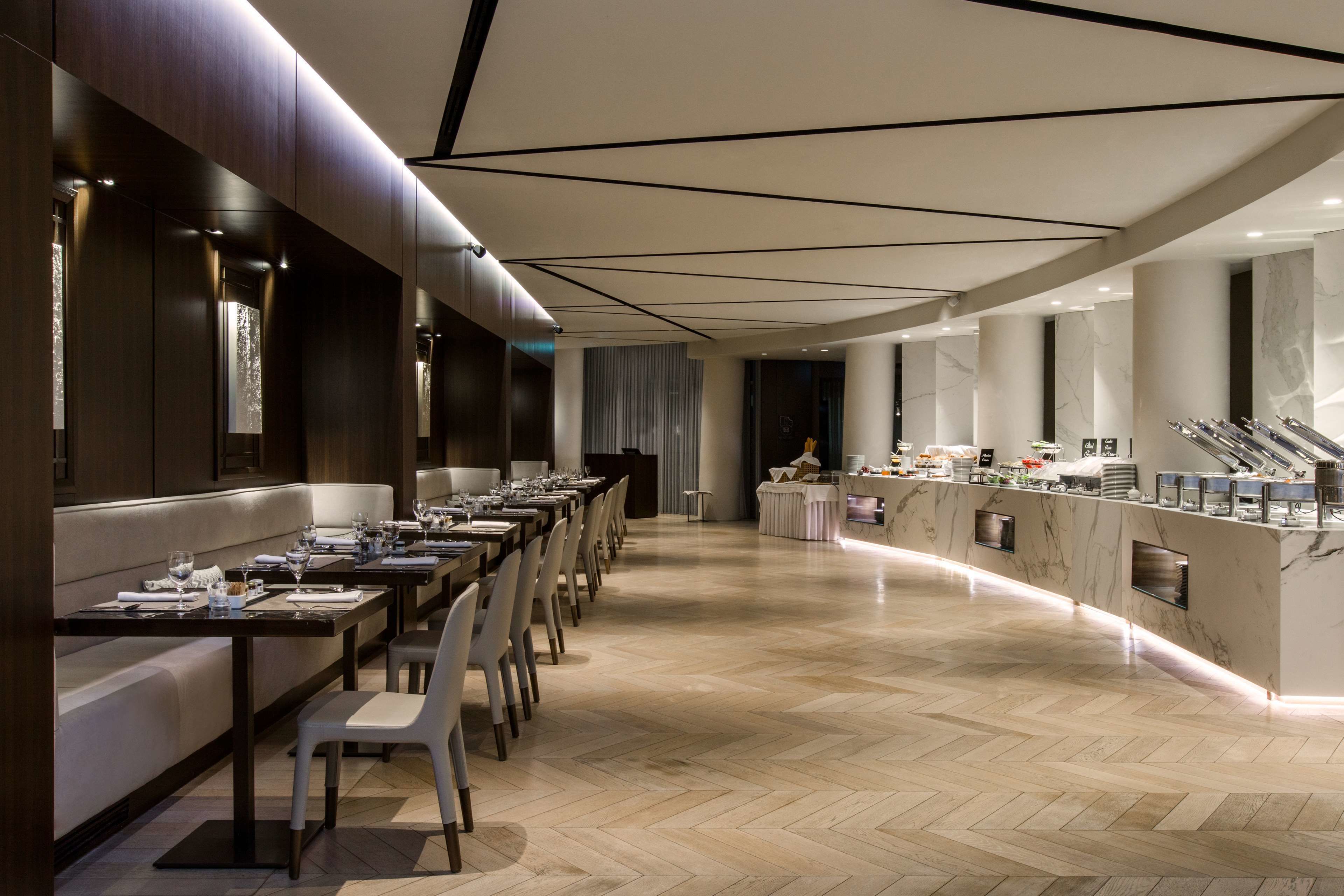 A modern restaurant with long dining tables, a marble counter, and a herringbone floor pattern.