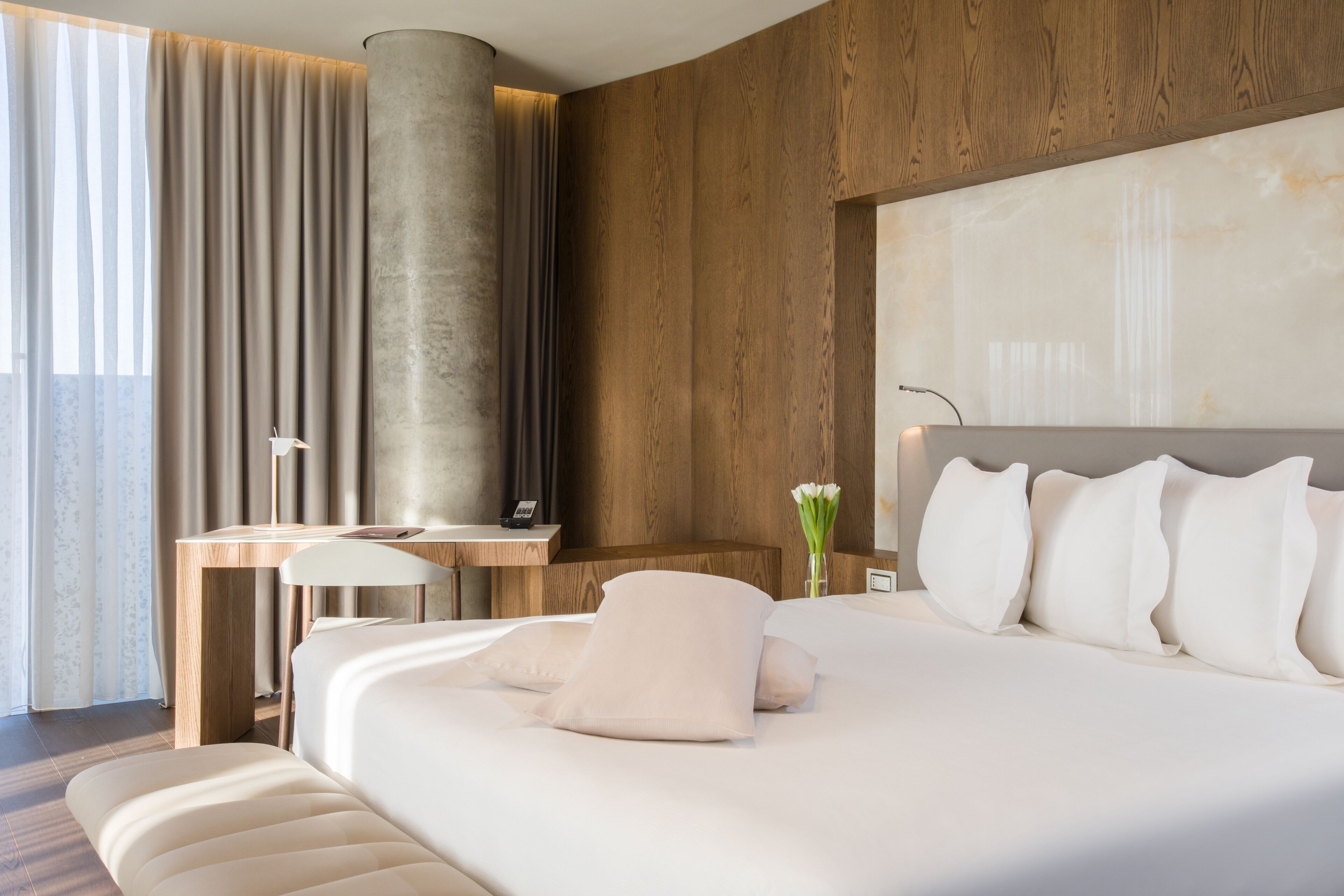 A modern hotel room with a large bed, wooden paneling, a desk with a lamp, and a view of the outdoors.