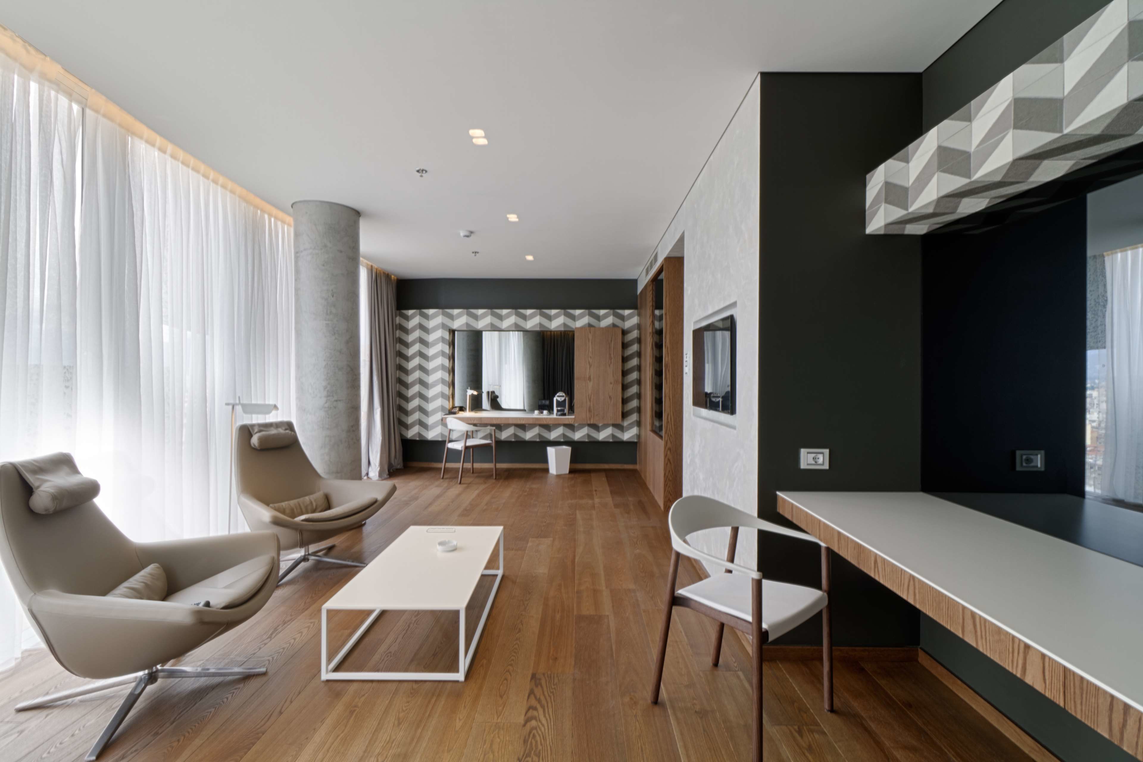 A modern living room with a wooden floor, a white coffee table, a beige sofa, and a white chair. There is a kitchen area with a herringbone-patterned backsplash and a window with sheer curtains.