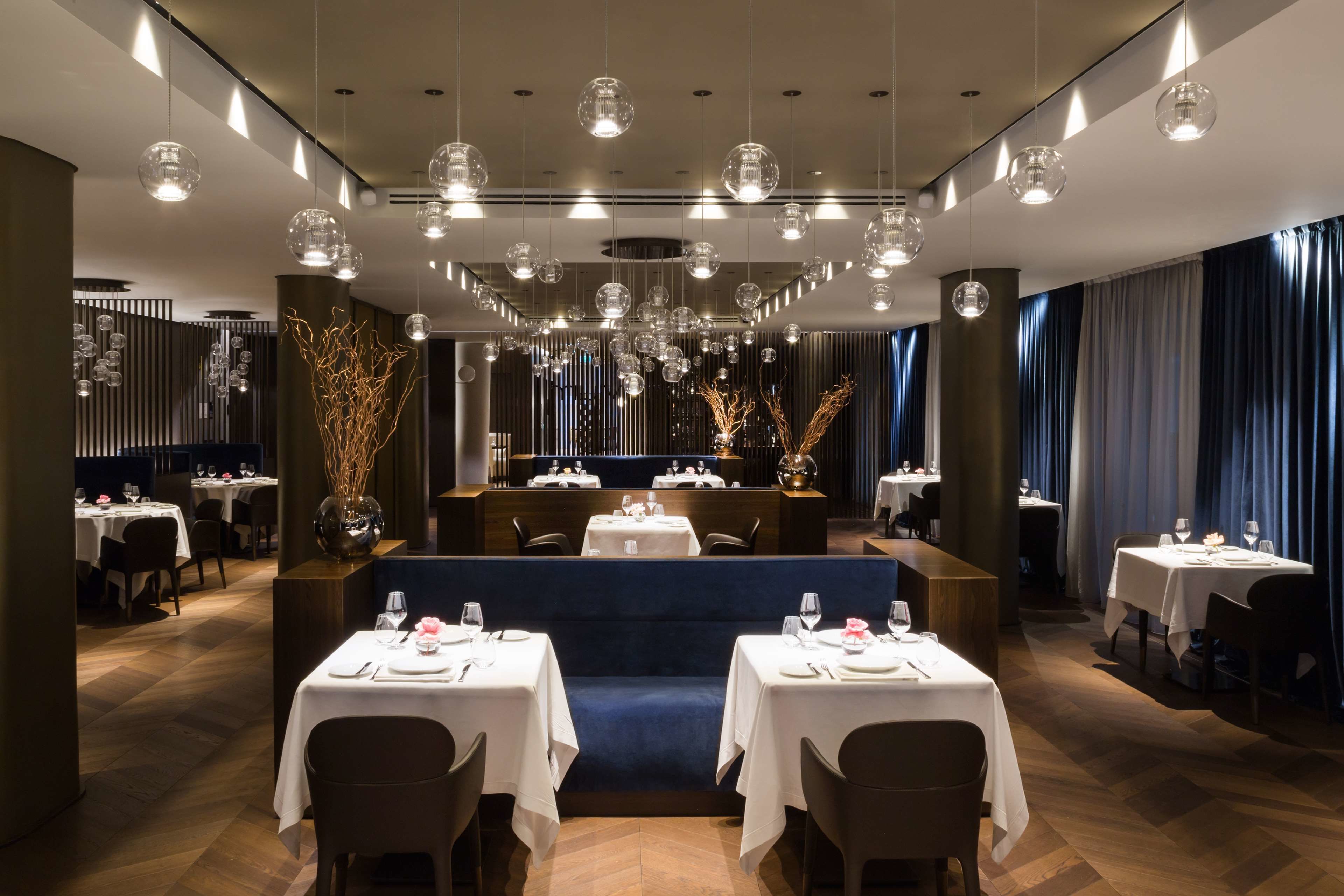 A modern restaurant interior with multiple dining tables, a central seating area, and hanging spherical light fixtures.