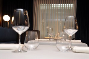 A dining table set with wine glasses and a candle, with a view of a cityscape through sheer curtains.