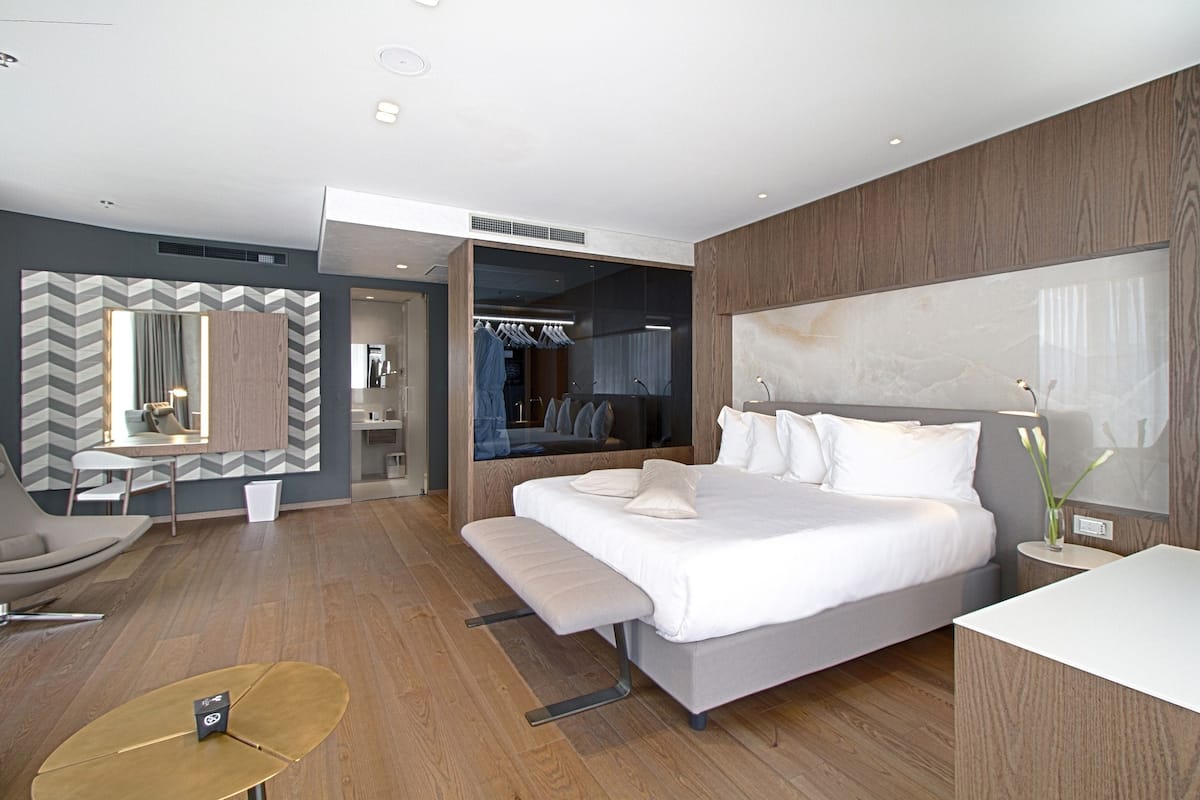 Una habitación de hotel moderna con una cama grande, una zona de estar con un banco, una pequeña mesa redonda y un espejo.
