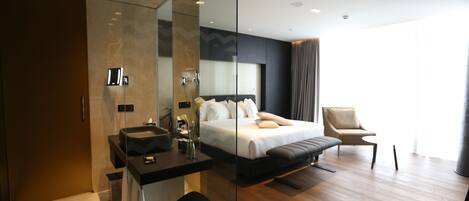 A modern hotel room with a glass partition, a large bed, a sitting area, and a bathroom with a sink and mirror.