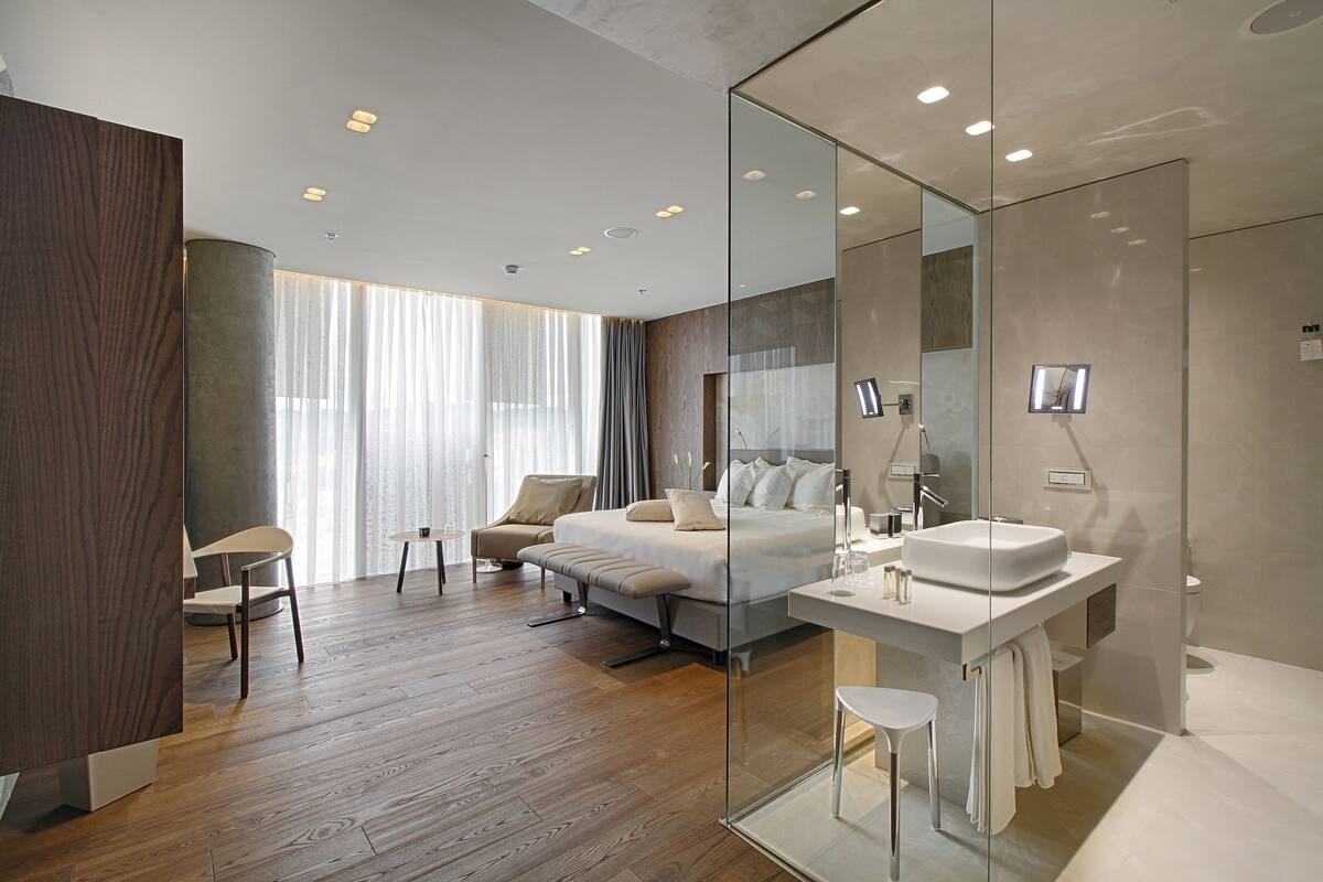 Una habitación de hotel moderna con piso de madera, una cama grande, un área de descanso con una silla y una mesita, un área de ducha con mampara de vidrio y un espejo grande.