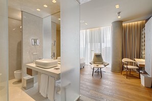 A modern bathroom with a large mirror, a sink, and a shower. There is a chair and a table in the adjacent room.