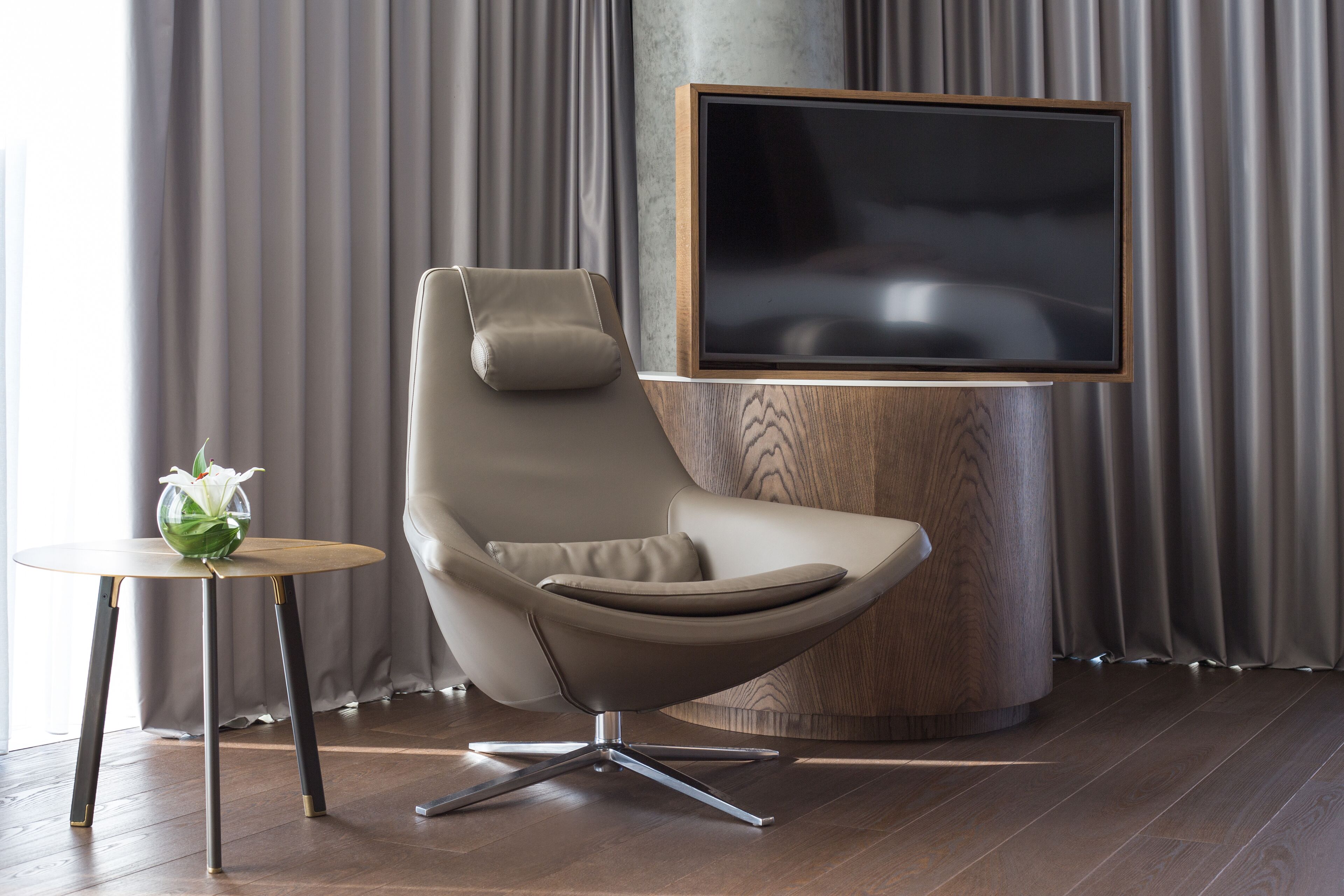 A modern living room with a leather armchair, a small round table with a vase of flowers, and a large flat-screen TV mounted on a wooden stand.