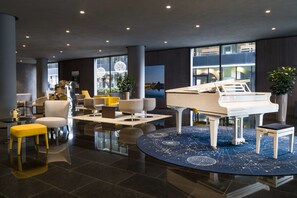 A modern lobby with a grand piano, comfortable seating, and large windows offering views of the city.