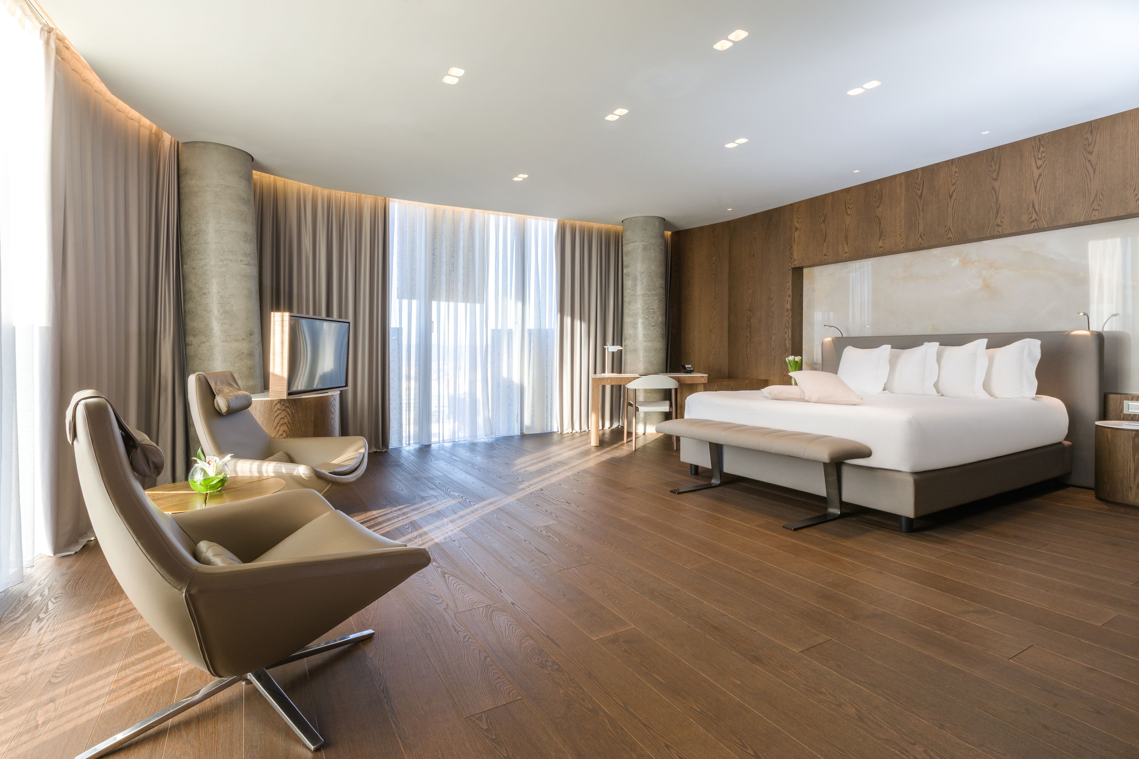 A modern hotel room with a large bed, a seating area with a chair and a small table, and a large window with curtains.