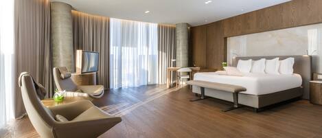 A modern hotel room with a large bed, a seating area with a chair and a small table, and a large window with curtains.