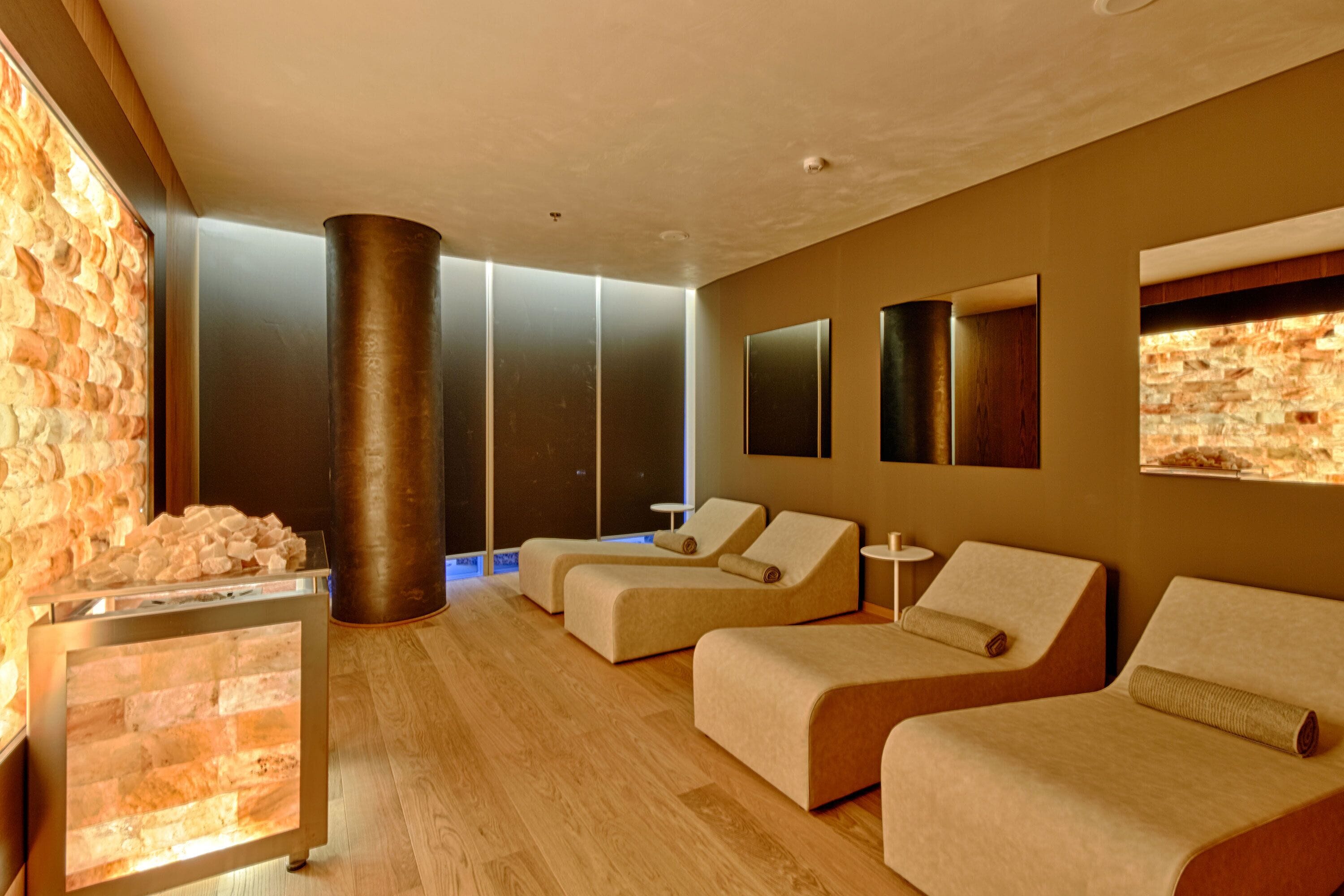 A modern spa room with a stone wall, a massage table, and two lounge chairs.
