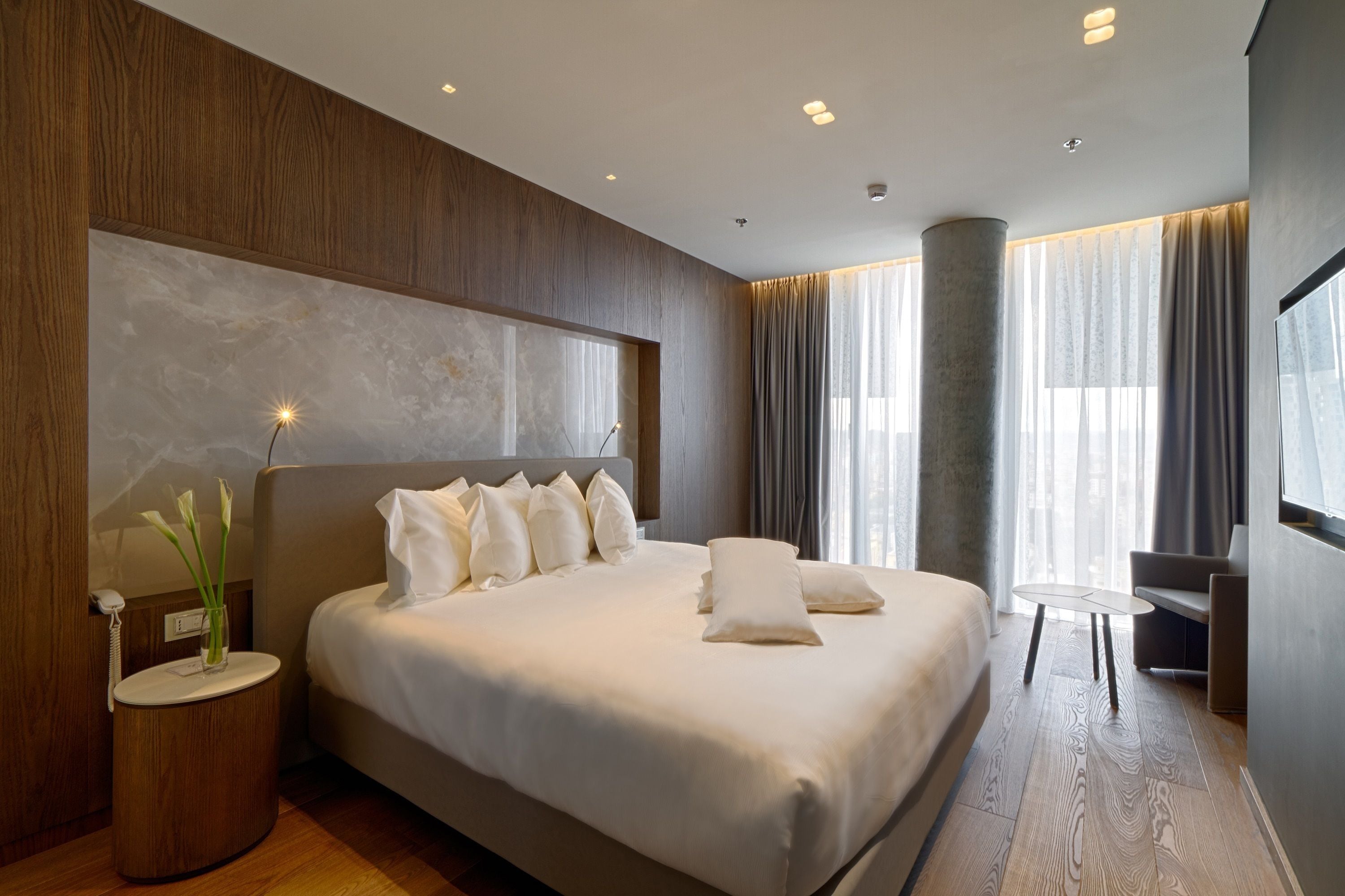 A modern hotel room with a large bed, a bedside table with a lamp, and a view of the city through large windows.