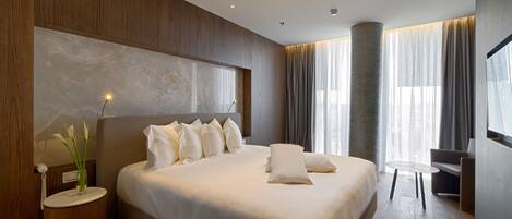 A modern hotel room with a large bed, a bedside table with a lamp, and a view of the city through large windows.