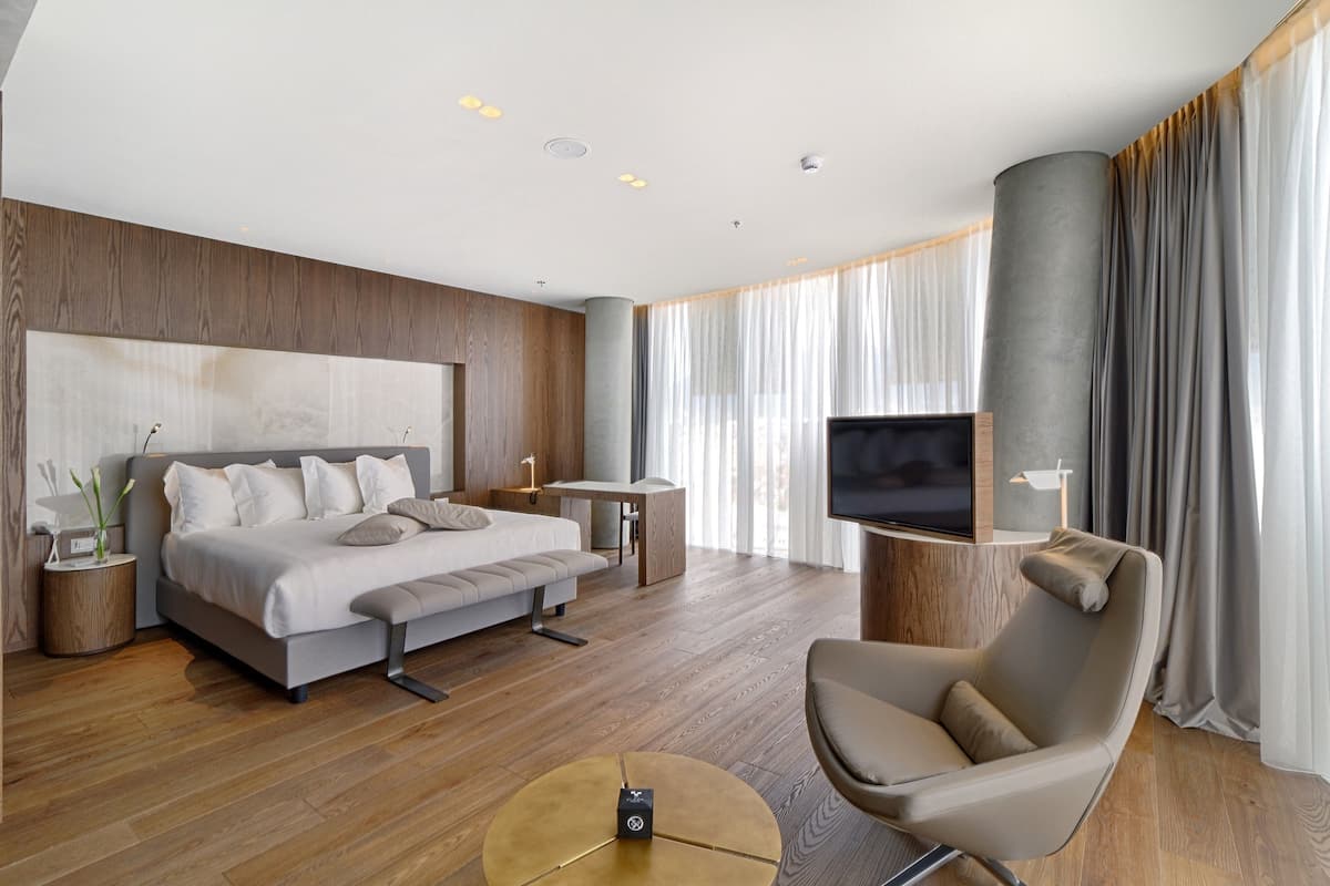 Una habitación de hotel moderna con una cama grande, televisión, zona de estar y suelo de madera.
