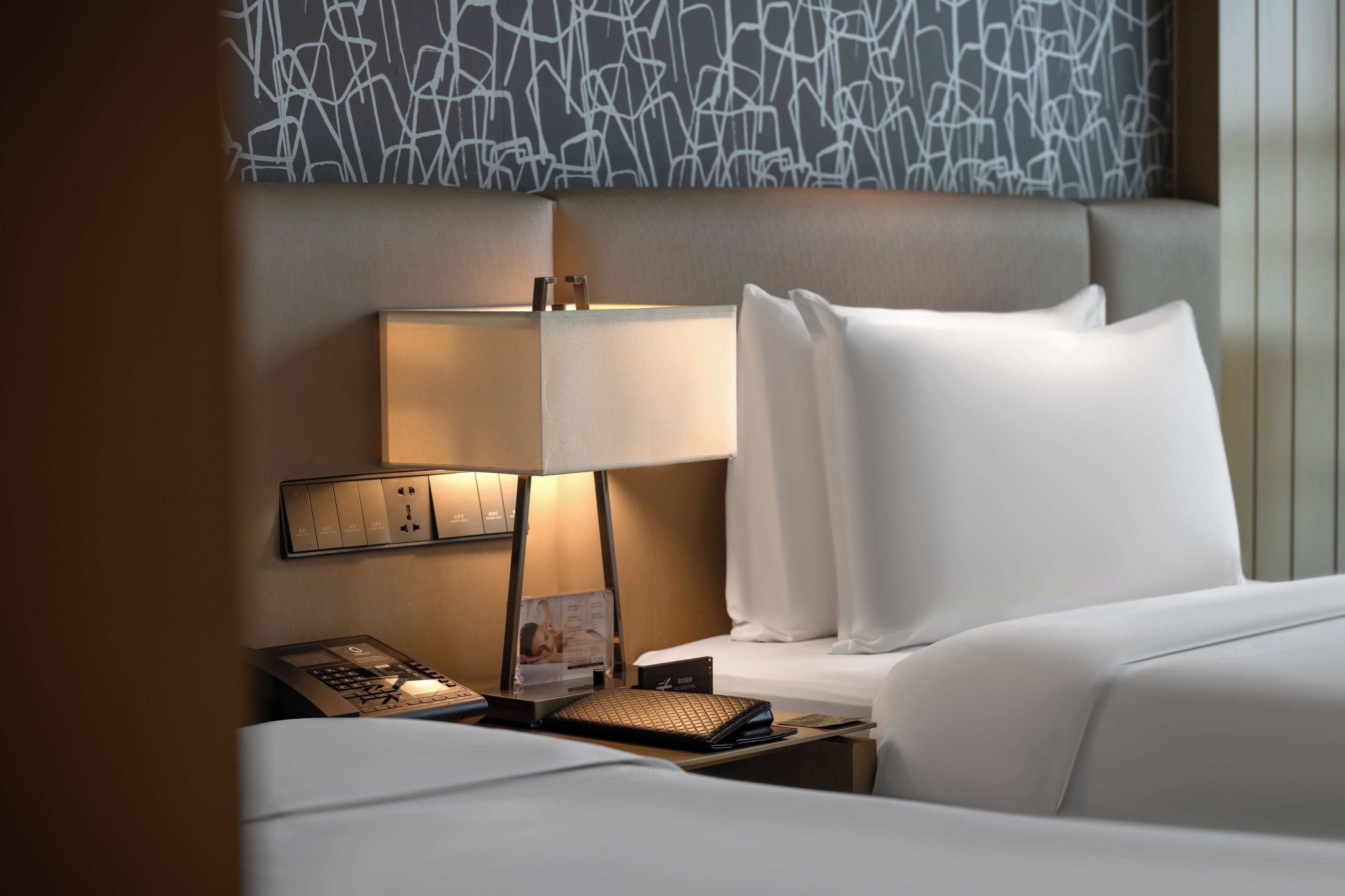 A hotel room with a bed, a bedside table with a lamp, a phone, and a picture frame.