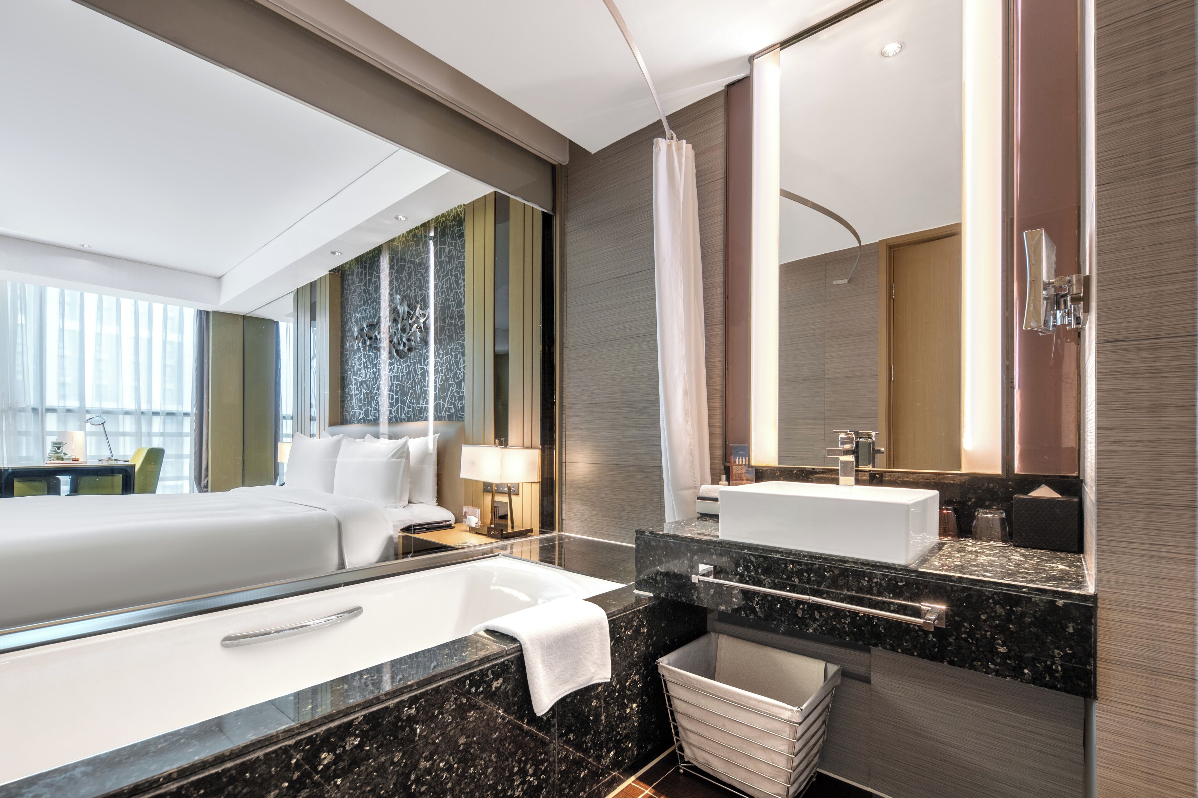 A modern hotel room with a large bed, a separate bathroom with a bathtub, a vanity with a mirror, and a large window offering a view.