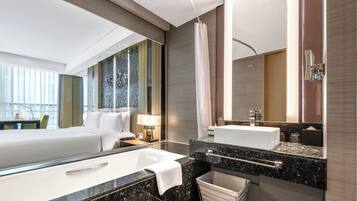 A modern hotel room with a large bed, a separate bathroom with a bathtub, a vanity with a mirror, and a large window offering a view.