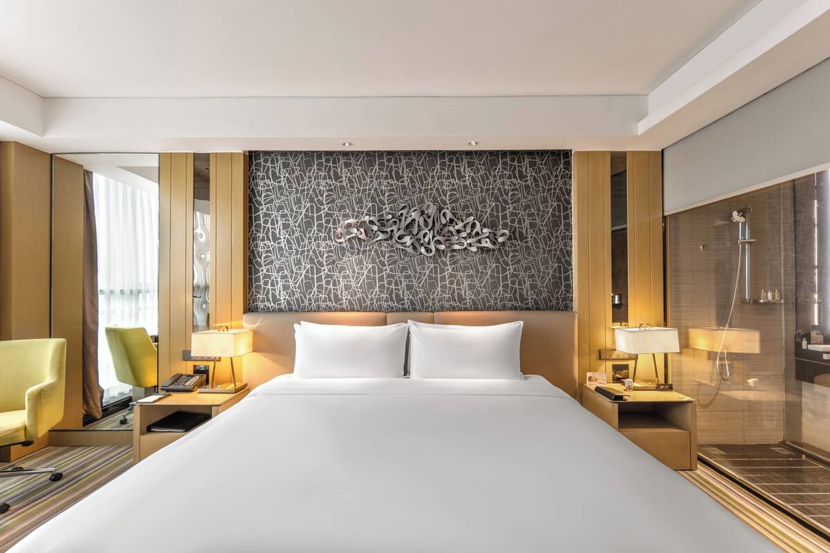 Une chambre d’hôtel avec un grand lit, deux tables de chevet avec des lampes, une chaise, une œuvre d’art murale et un espace douche.