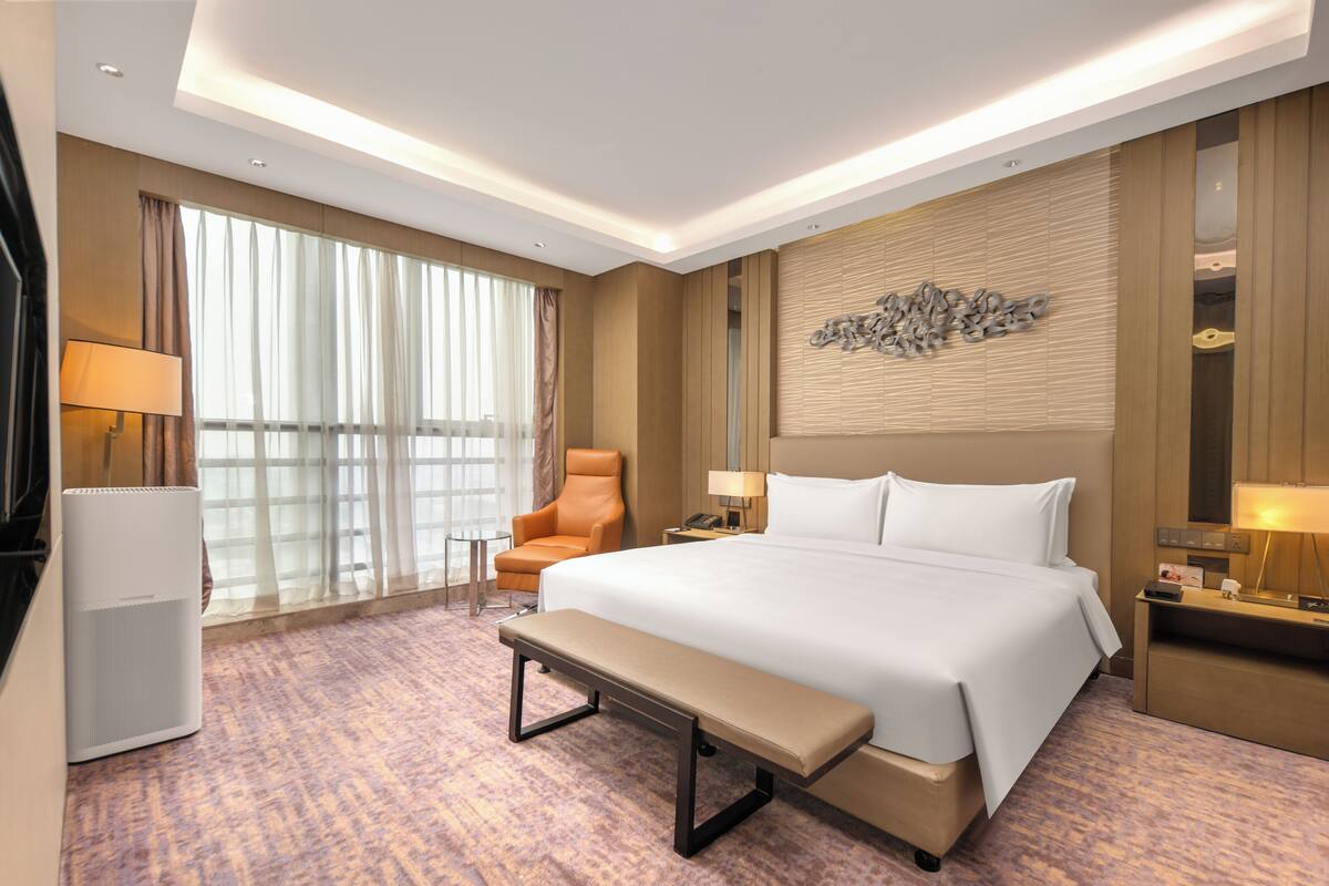 Une chambre d’hôtel avec un grand lit, un banc, une chaise, un bureau, une lampe, une télévision et une fenêtre avec des rideaux.