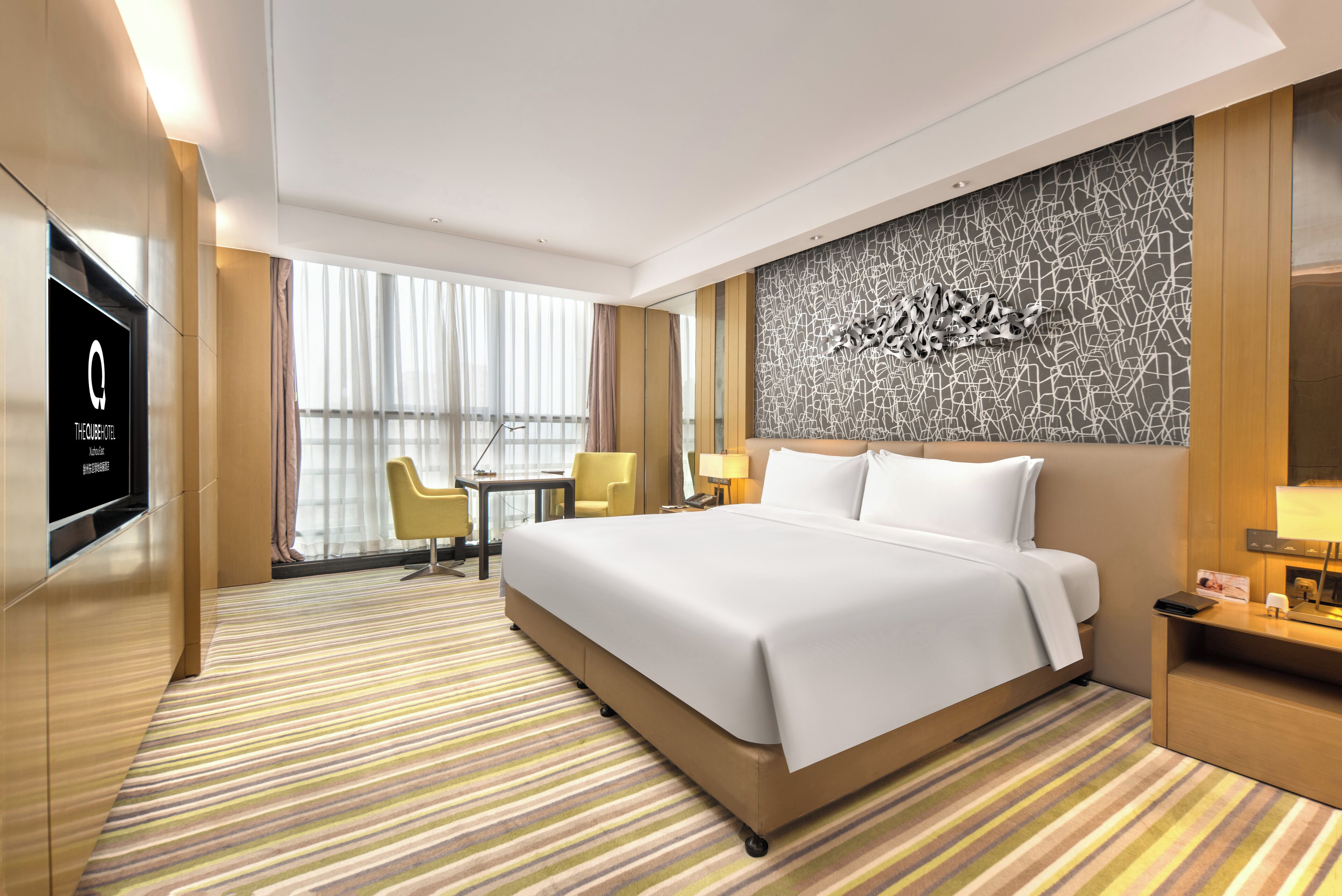 A modern hotel room with a large bed, a flat-screen TV, a desk with a chair, and a decorative wall.