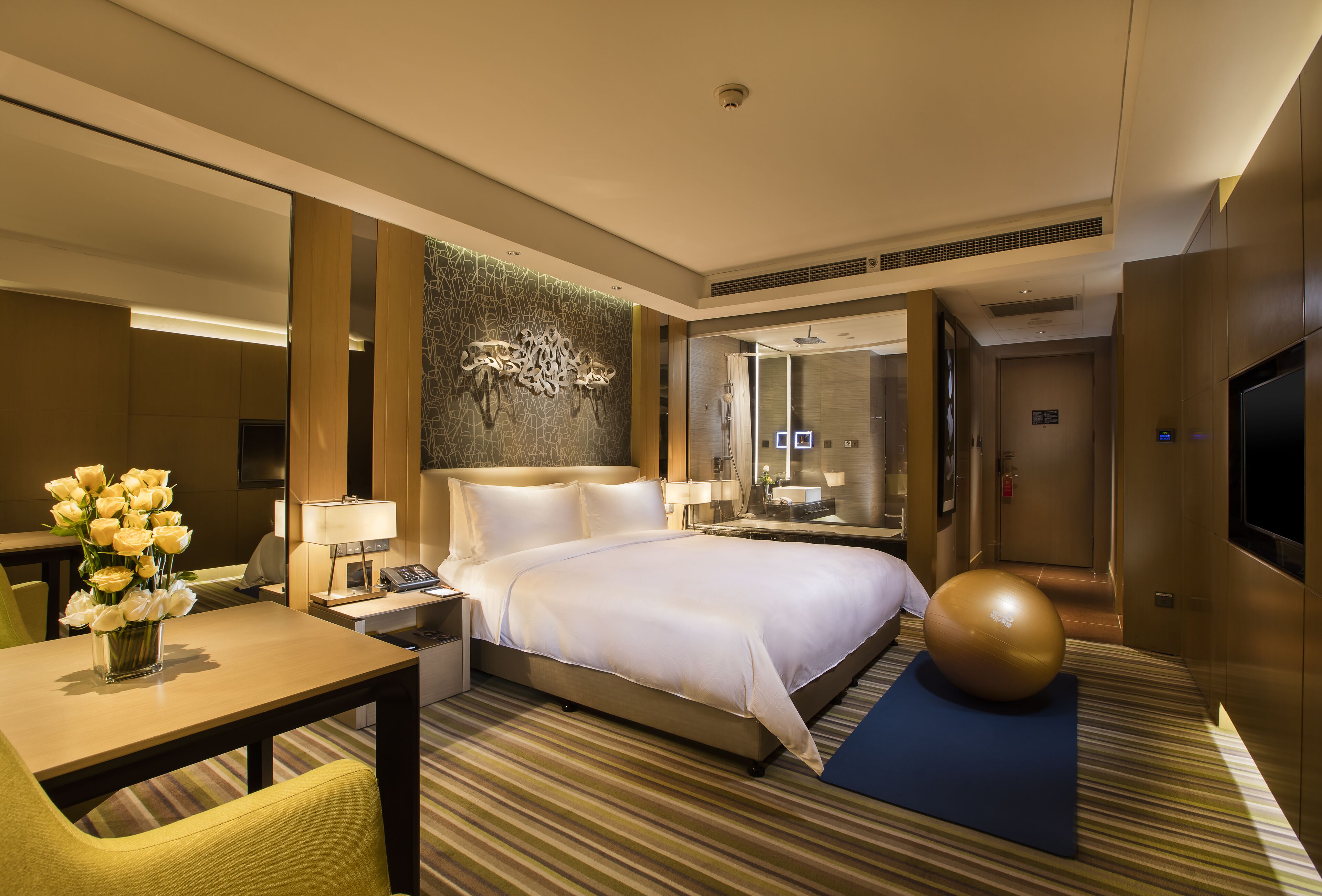 Habitación de hotel con una cama grande, un escritorio con lámpara, un florero y una pelota de ejercicios grande.