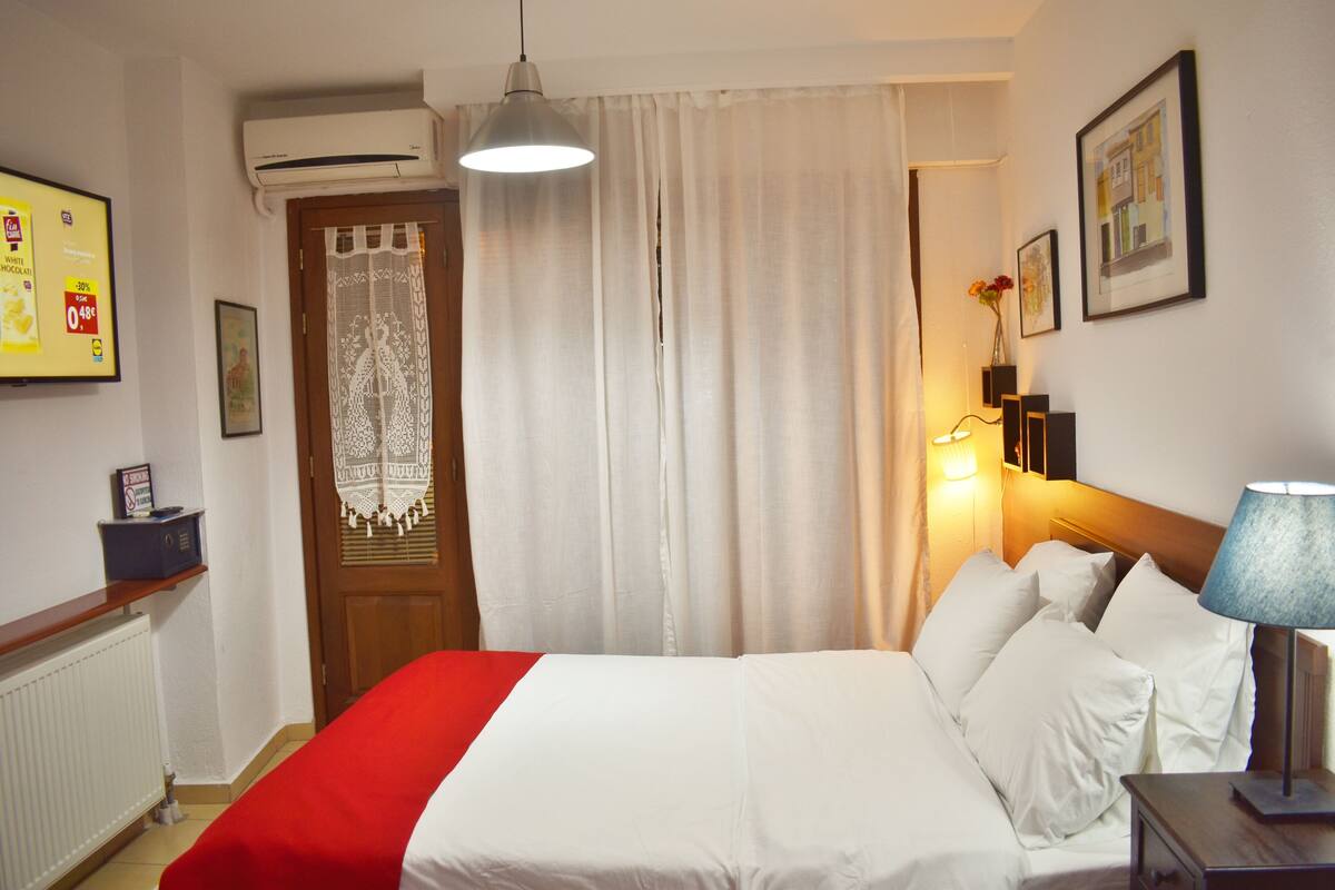 Ein Hotelzimmer mit einem Bett, einem Nachttisch, einer Lampe, einem Fenster mit Vorhängen, einer Wandklimaanlage und einem gerahmten Bild.