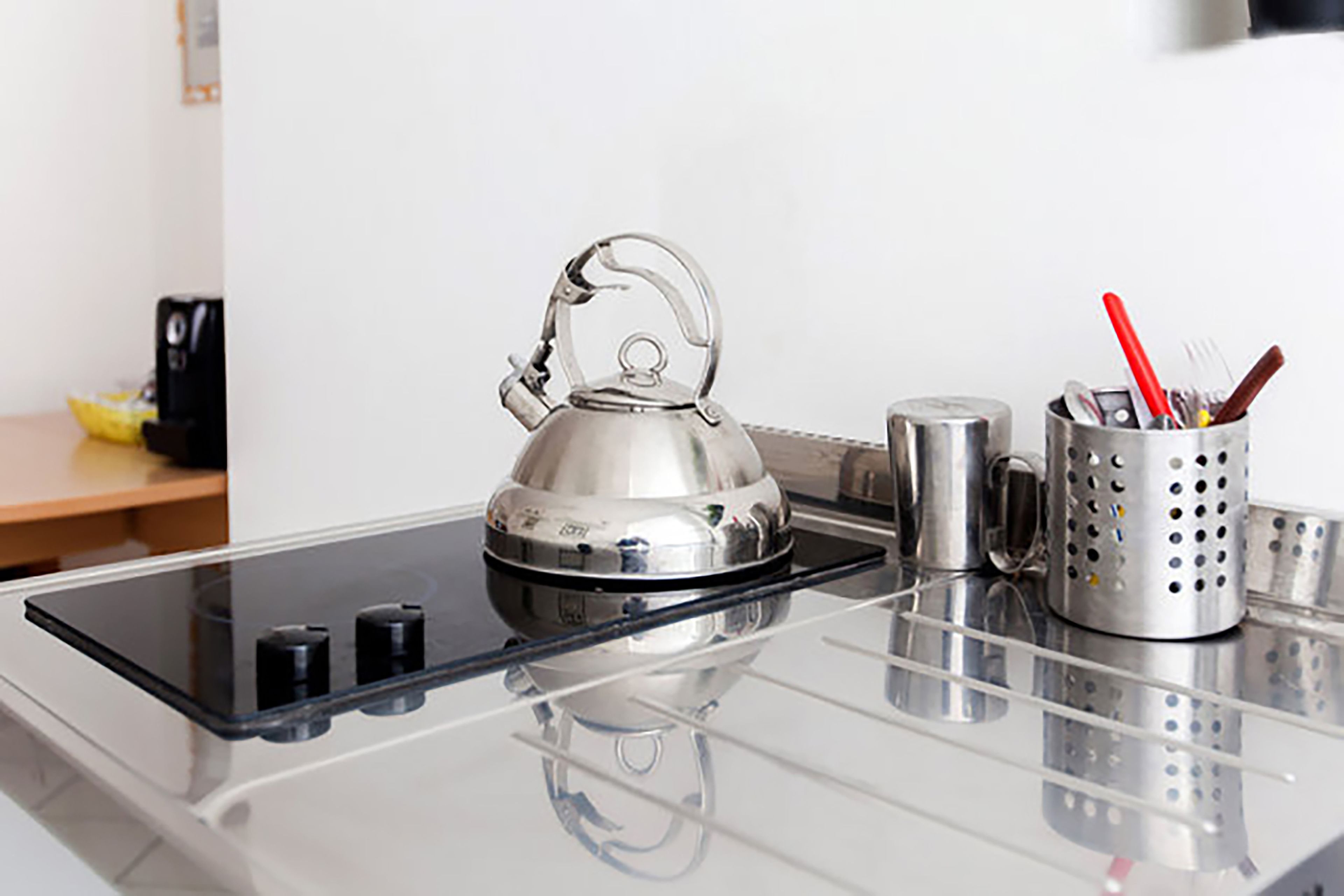 A stainless steel kettle on a glass-top stove, a stainless steel mug, and a utensil holder with a red spoon and fork.