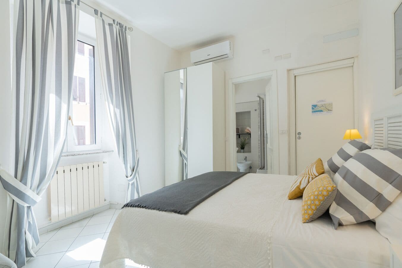 Een slaapkamer met een bed, gordijnen, een airconditioning, een deur en een kleine tafel met een lamp.