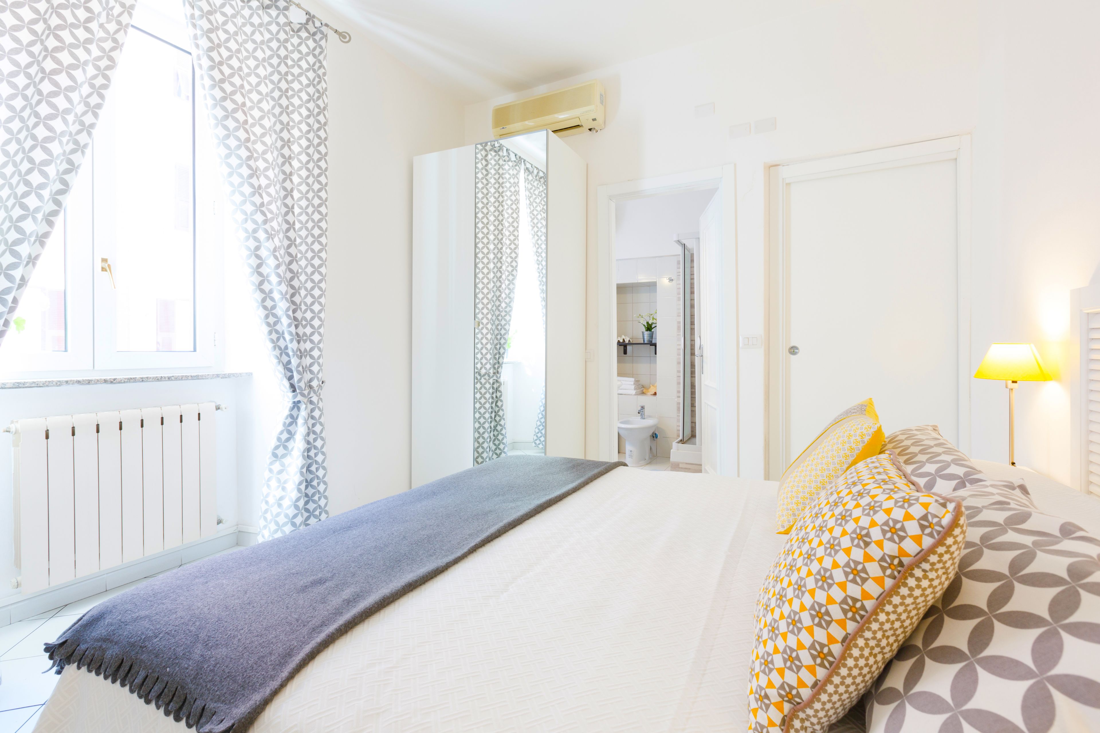 Un dormitorio con cama, lámpara de noche, radiador y baño visible a través de una puerta abierta.