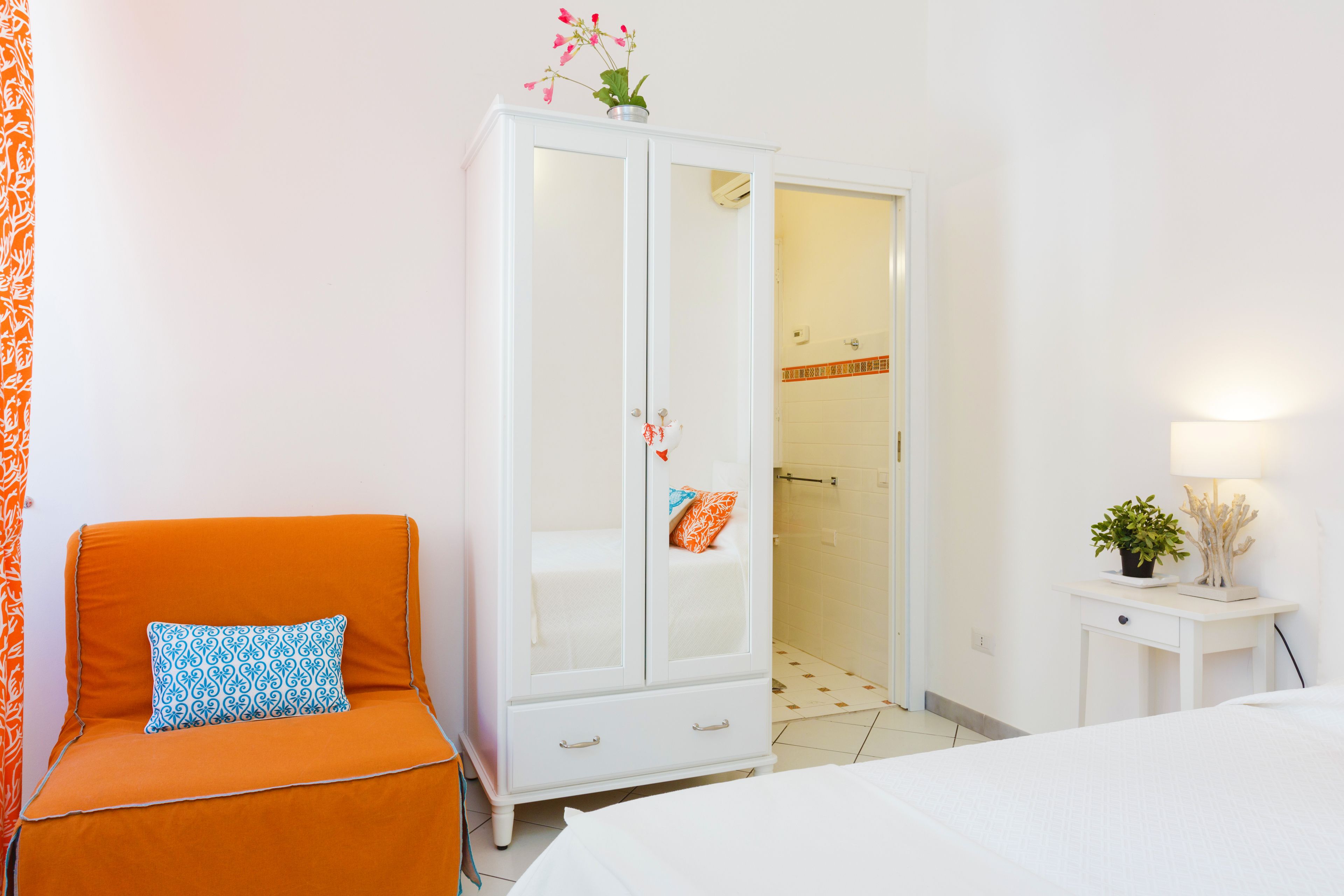 A bedroom with a bed, an orange chair with a patterned pillow, a white wardrobe, and a bedside table with a lamp and a plant.