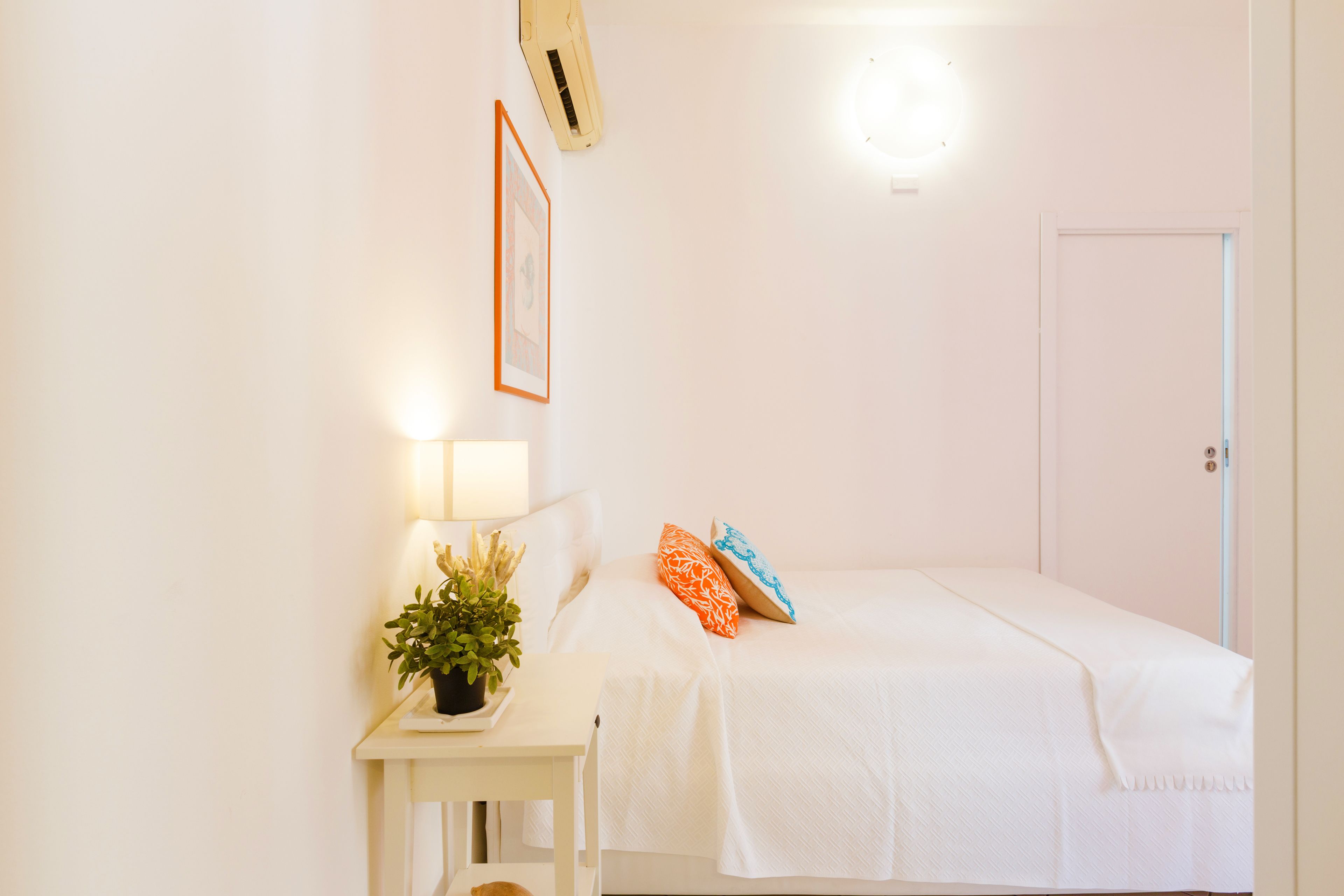 Habitación blanca con una cama, una mesita de noche con lámpara y una planta en maceta.