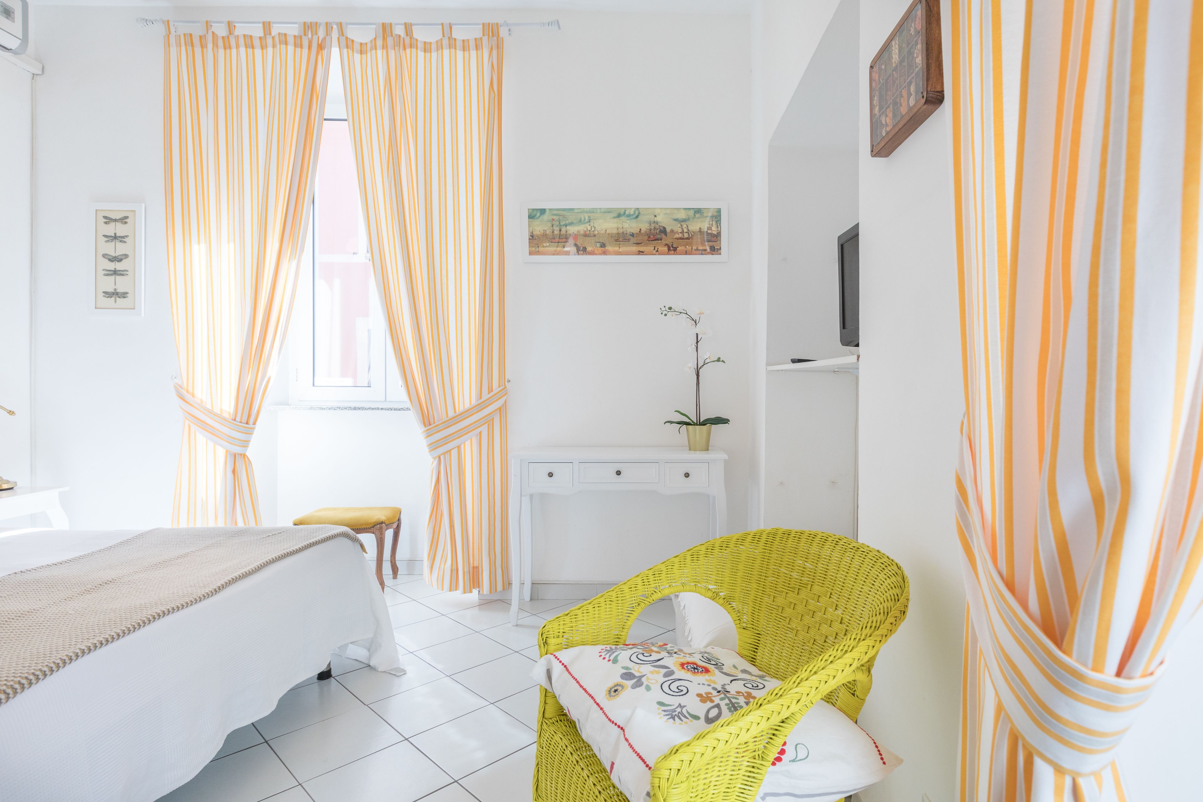 Una habitación de hotel con una cama, un sillón de mimbre amarillo, un escritorio blanco y cortinas a rayas.