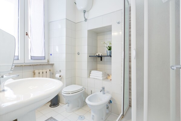 A modern bathroom with a sink, toilet, and bidet. There is a window with a curtain, a towel rack, and a shelf with toiletries.