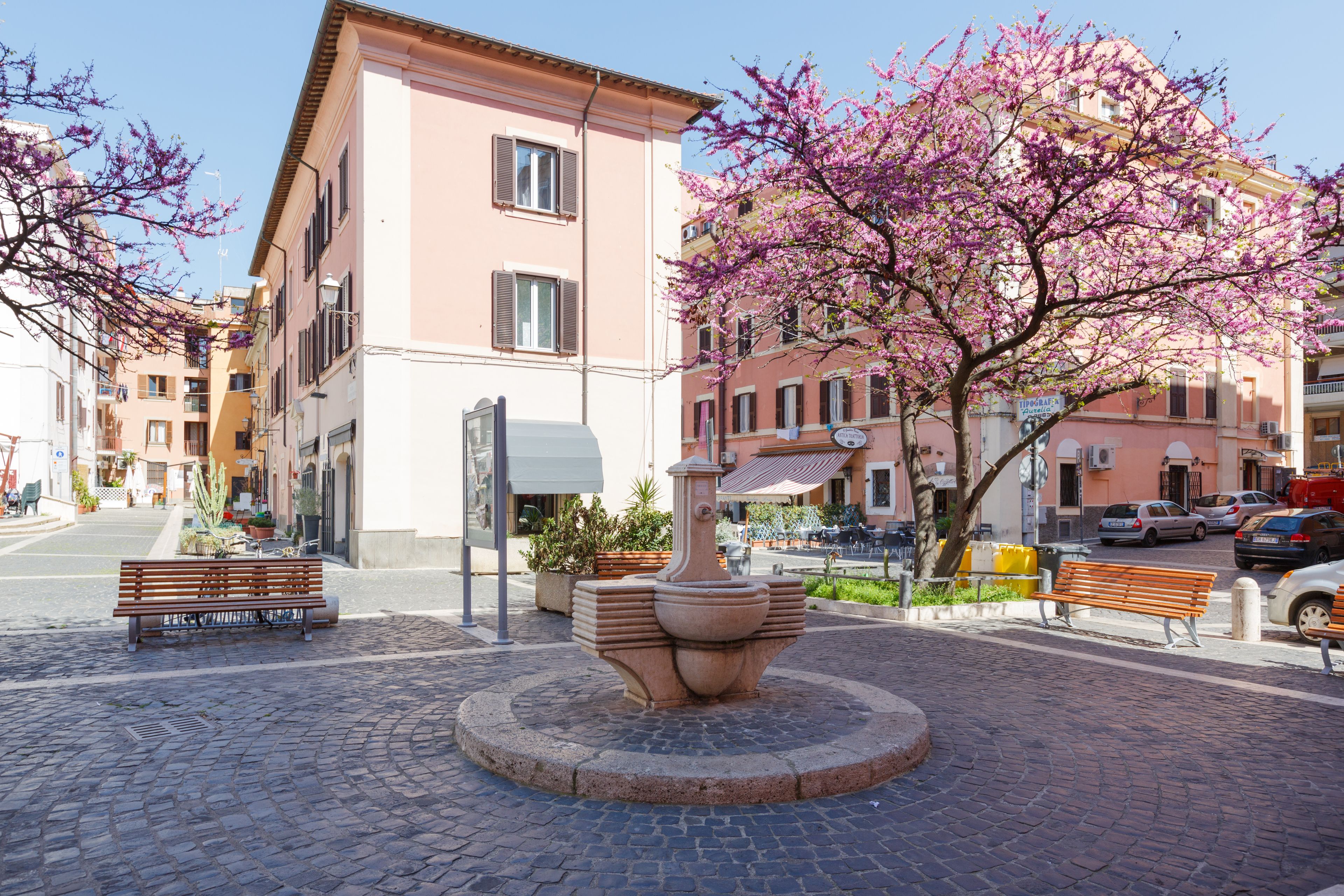 Une place pavée avec une fontaine centrale, des bancs et un bâtiment rose doté de balcons.