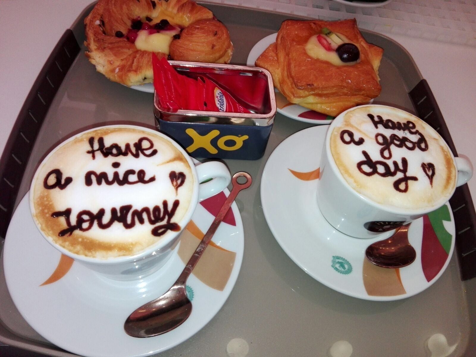 Two cups of coffee with motivational messages, a croissant, a pastry with fruit, and a box of chocolates on a tray.