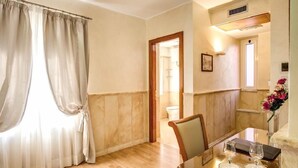 A hotel room with a wooden floor, a dining table with two glasses, a chair, a window with curtains, and a door leading to the bathroom. - Pantheon Rooms (Rome)