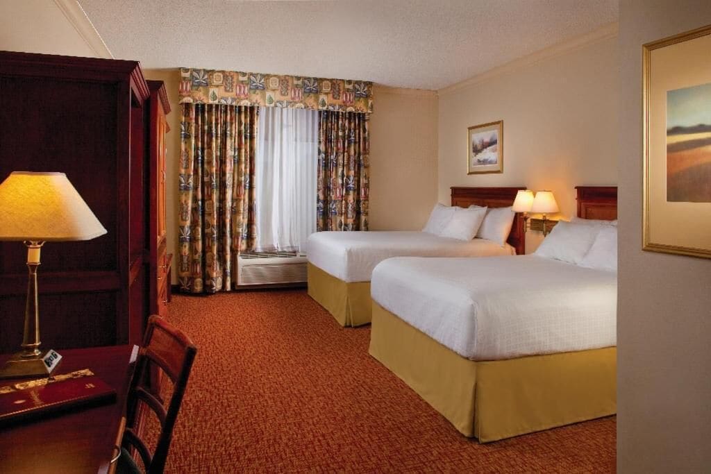 Habitación de hotel con dos camas, un escritorio con lámpara, una silla, una ventana con cortinas y un cuadro en la pared.