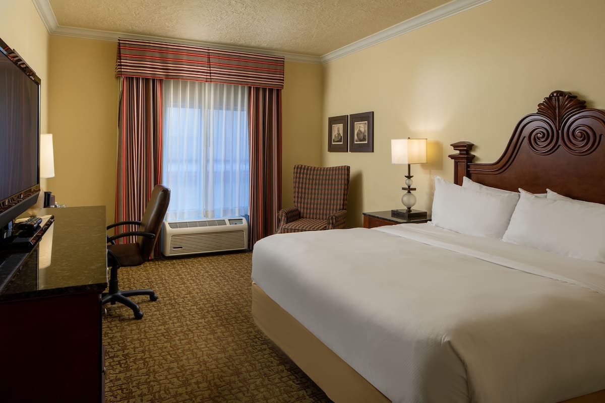 A hotel room with a large bed, a desk with a computer, a chair, an air conditioning unit, and a window with curtains.