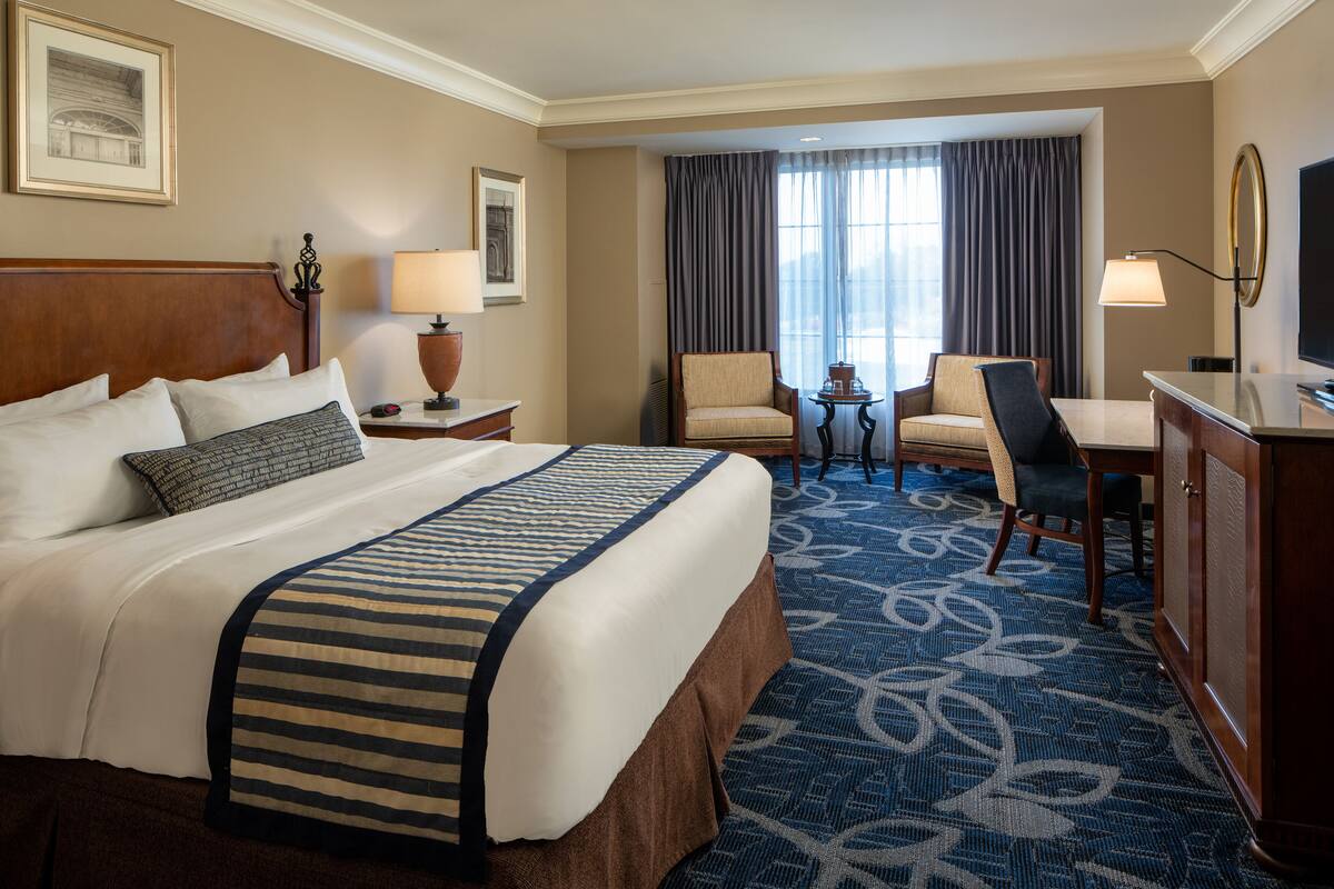 A hotel room with a large bed, two chairs, a desk, a TV, and a window with curtains.