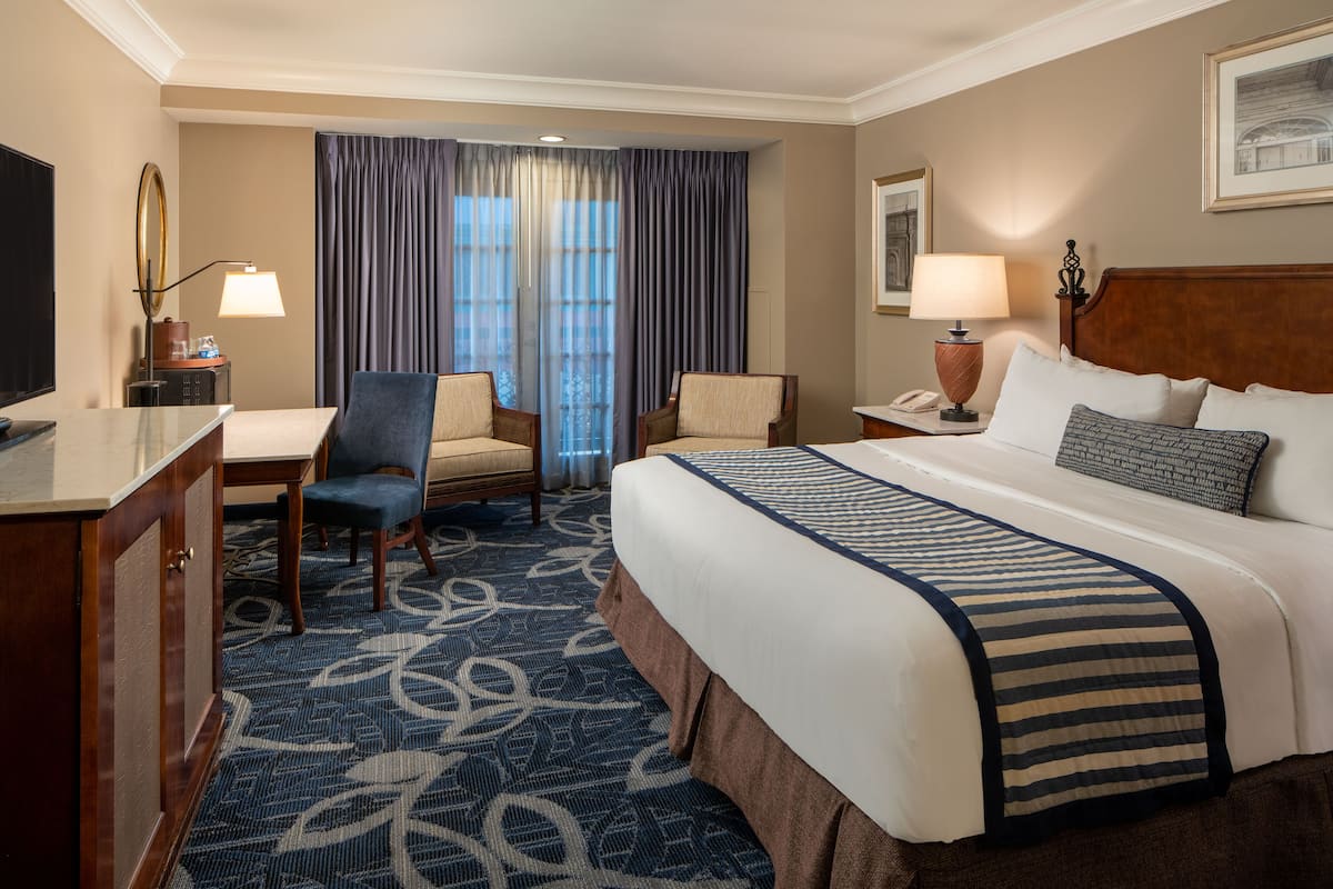 A hotel room with a large bed, two armchairs, a desk, a television, and a window with curtains.